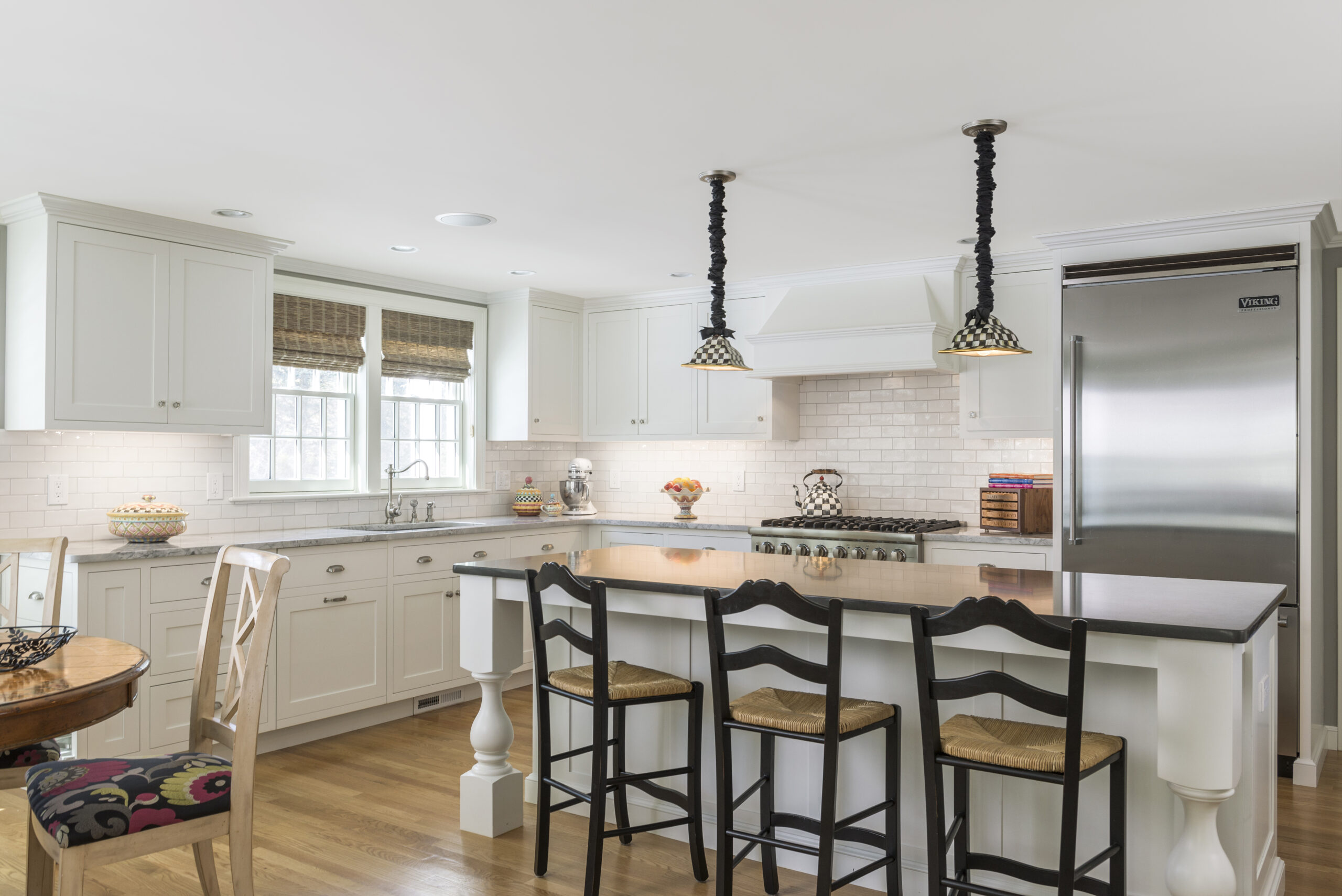 Modern kitchen with white cabinets, stainless steel appliances, a large island with black countertop, three barstools, wooden floor, and pendant lights—an example of Luxury Interior Design by an Interior Designer Massachusetts.