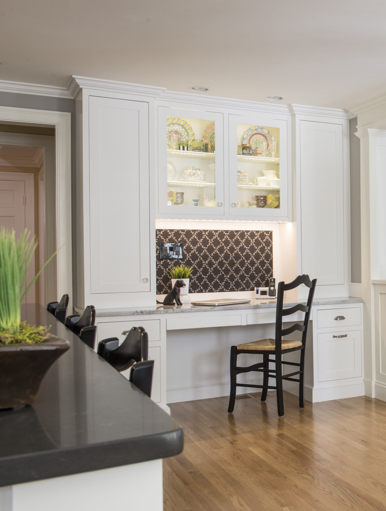 A built-in white desk with a black chair is set into a kitchen wall, featuring glass-front cabinets, open shelves, and a black patterned backsplash above a wooden floor—an elegant touch inspired by luxury interior design.
