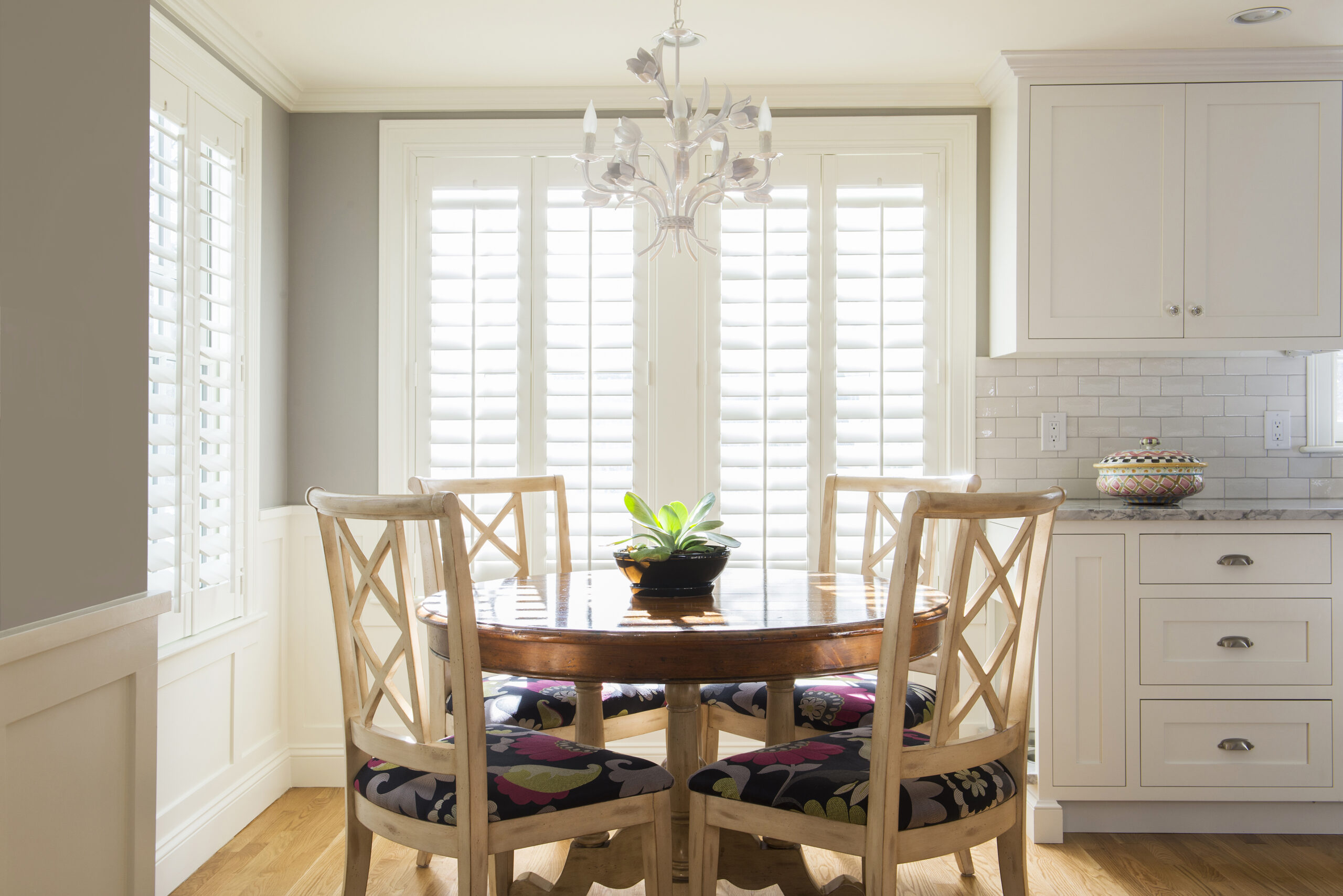 A round wooden dining table with four chairs sits in a bright kitchen nook, beautifully styled by an Interior Designer Massachusetts, featuring large shuttered windows, a white chandelier, and a potted plant centerpiece.