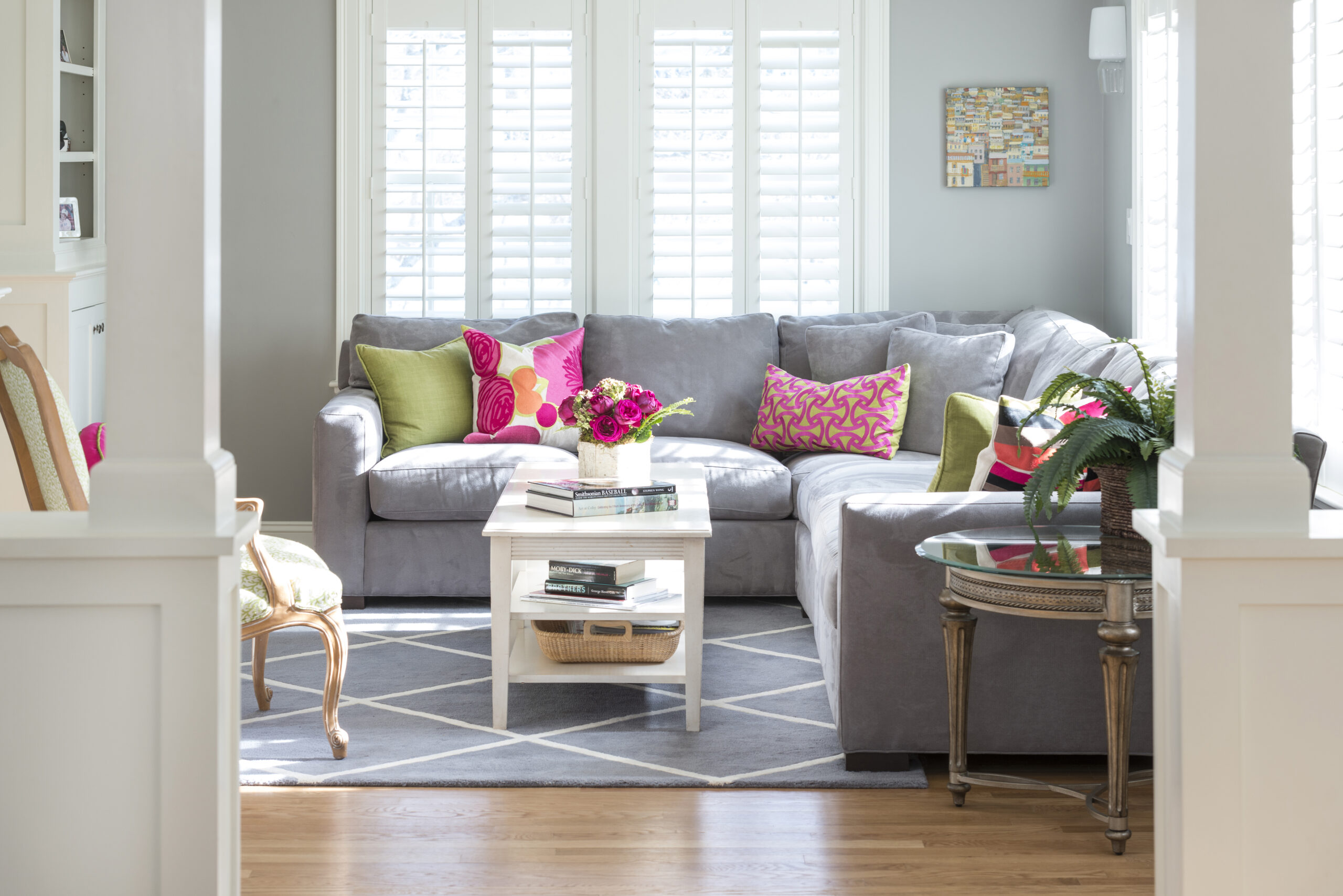 A bright living room with a gray sectional sofa, colorful pillows, a white coffee table with flowers, and large windows with white shutters showcases luxury interior design by an expert interior designer in Massachusetts.