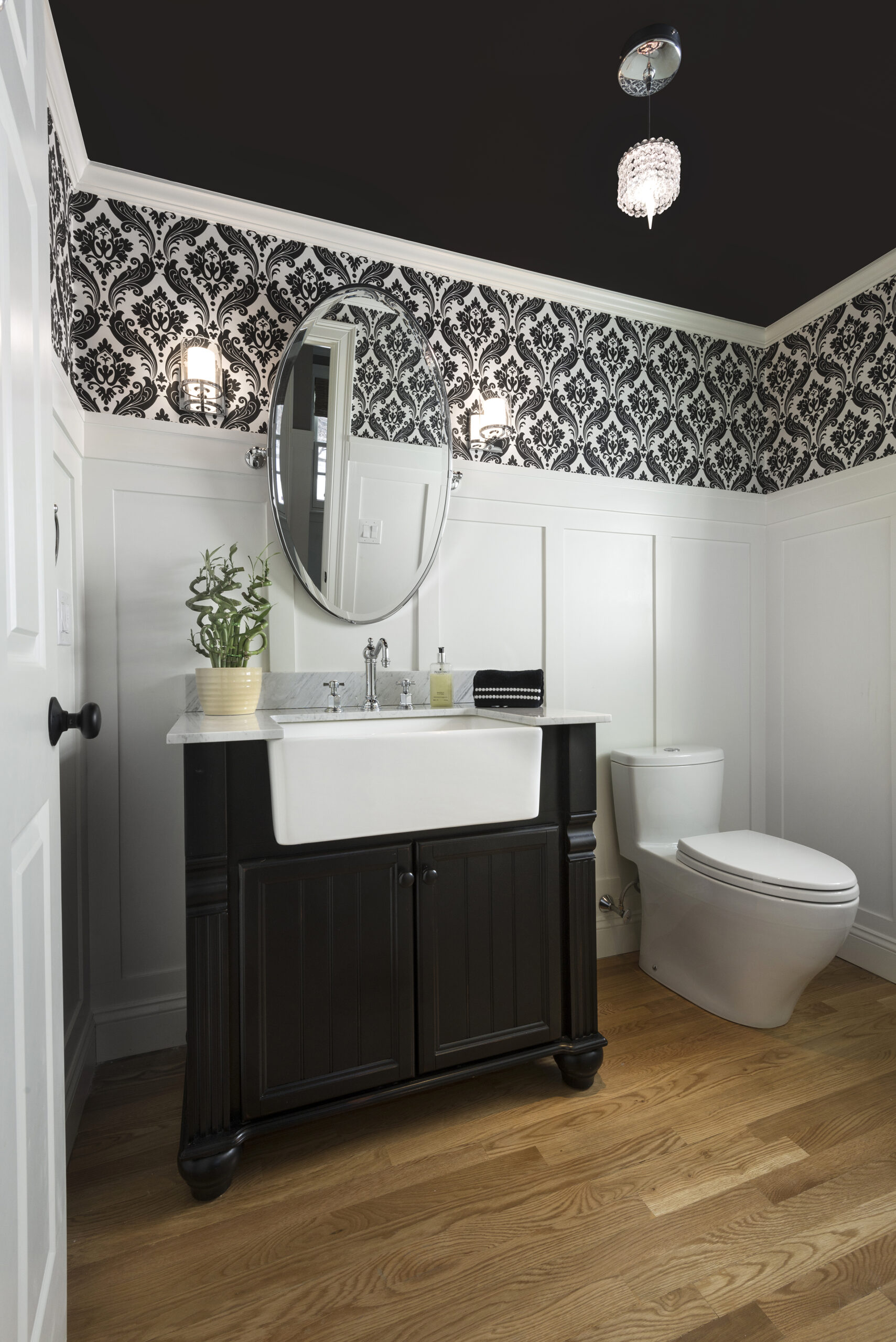 A bathroom with a black vanity, oval mirror, white sink, modern toilet, wood floor, and black and white wallpaper showcases luxury interior design by an expert interior designer in Massachusetts.