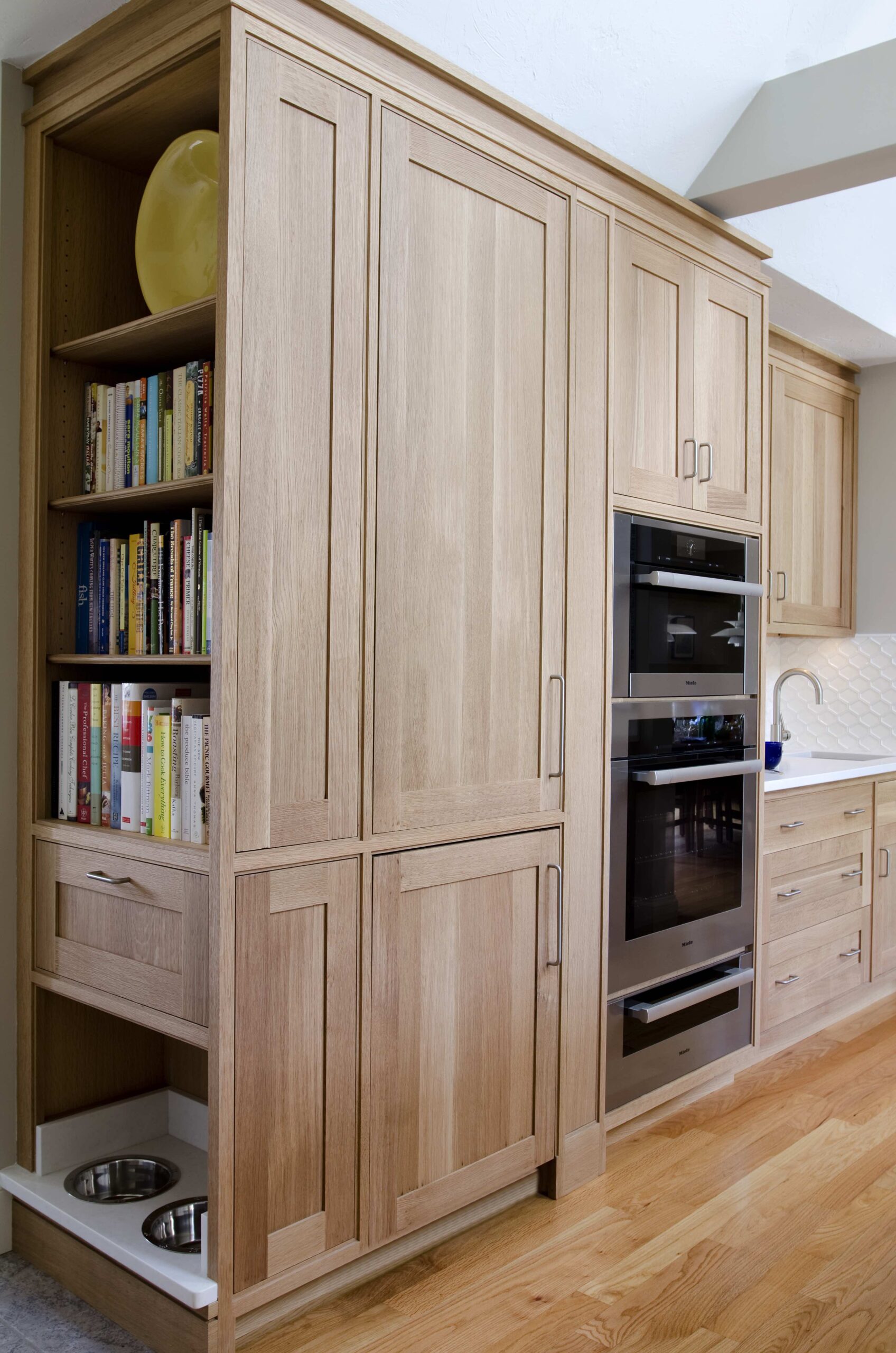 Tall wooden kitchen cabinets with built-in bookshelves, pull-out drawer, double wall ovens, and a pull-out tray with two stainless steel pet bowls on hardwood floors—a touch of luxury interior design by an interior designer in Massachusetts.