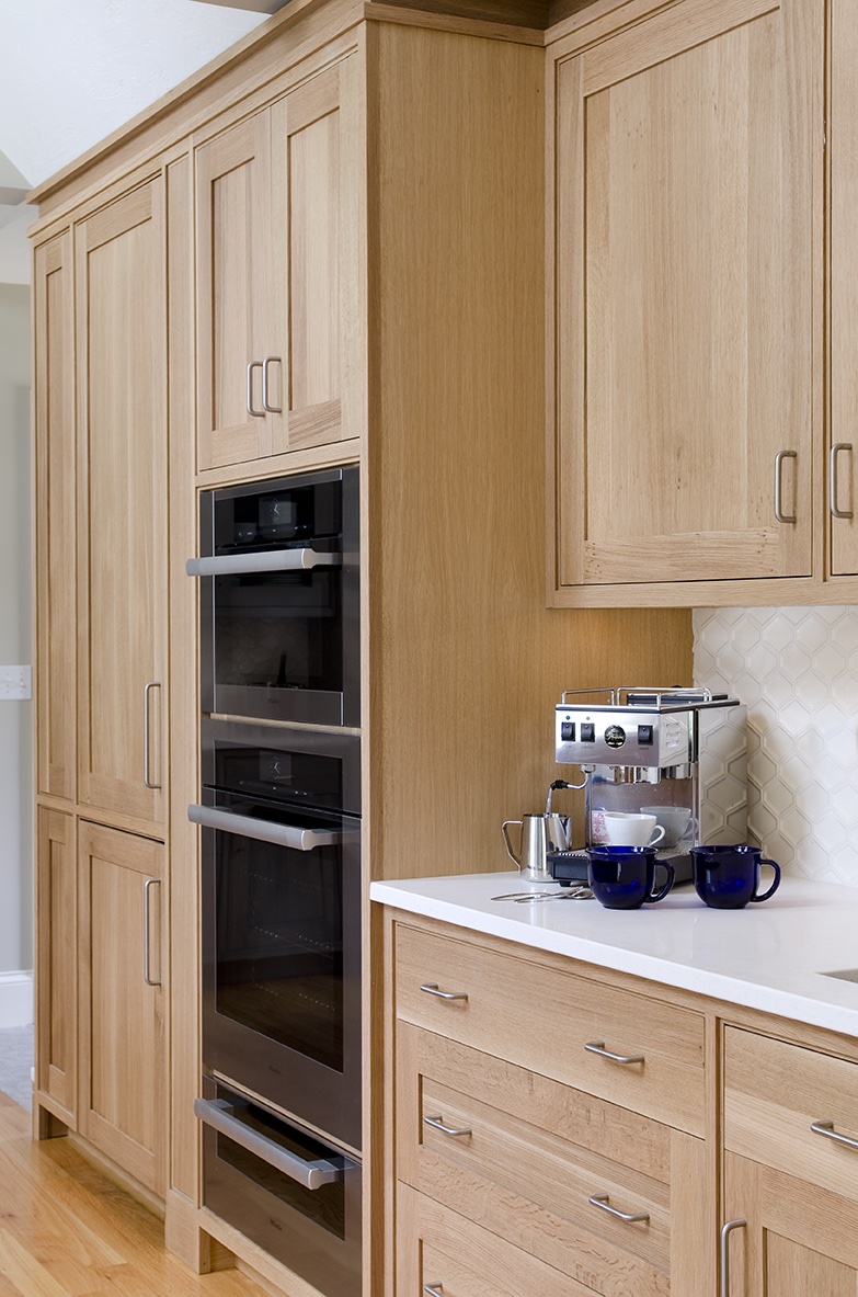 Light wood kitchen with built-in double oven, espresso machine, mugs, and white countertop. Geometric tile backsplash and floor-to-ceiling cabinetry highlight the luxury interior design by a top Interior Designer Massachusetts.