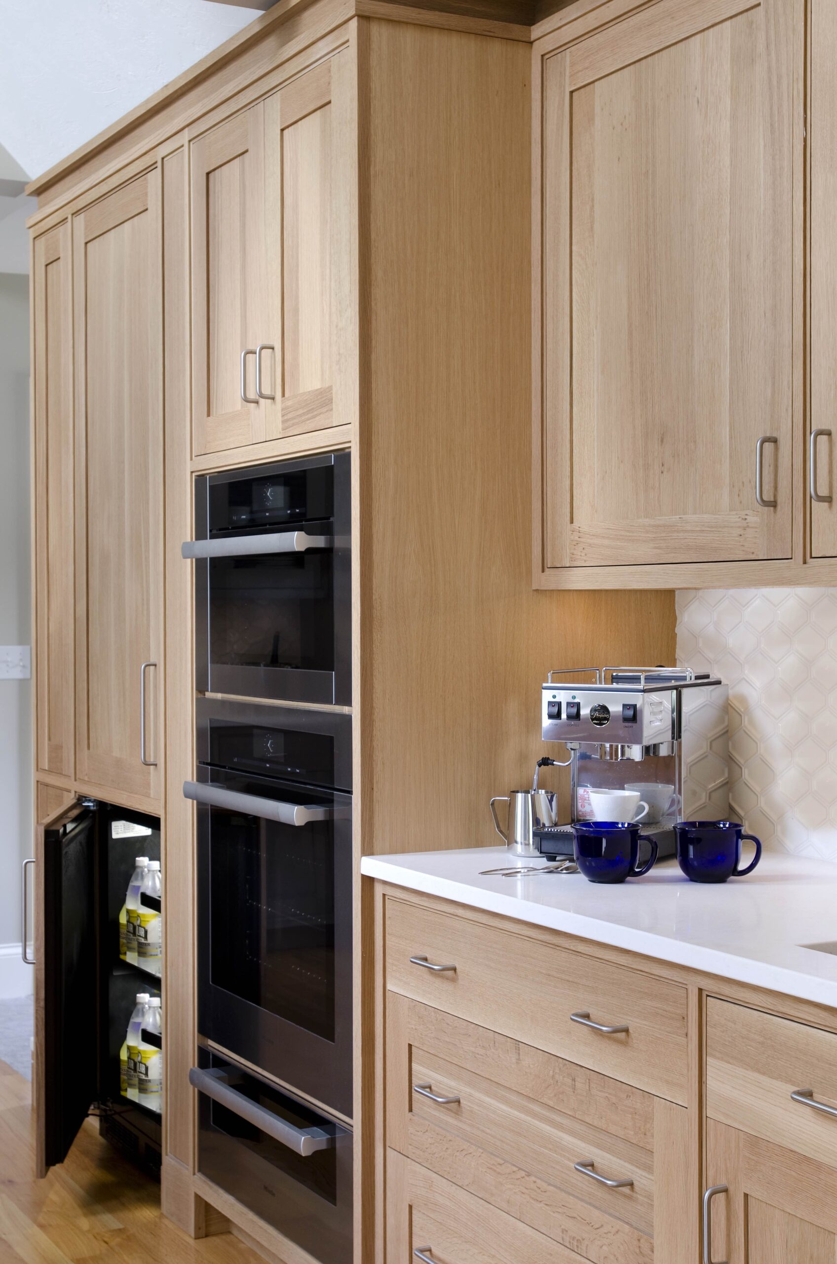 Modern kitchen with light wood cabinets, built-in double oven, partially open beverage fridge, and a countertop espresso machine with two blue mugs—an elegant example of luxury interior design by an interior designer in Massachusetts.