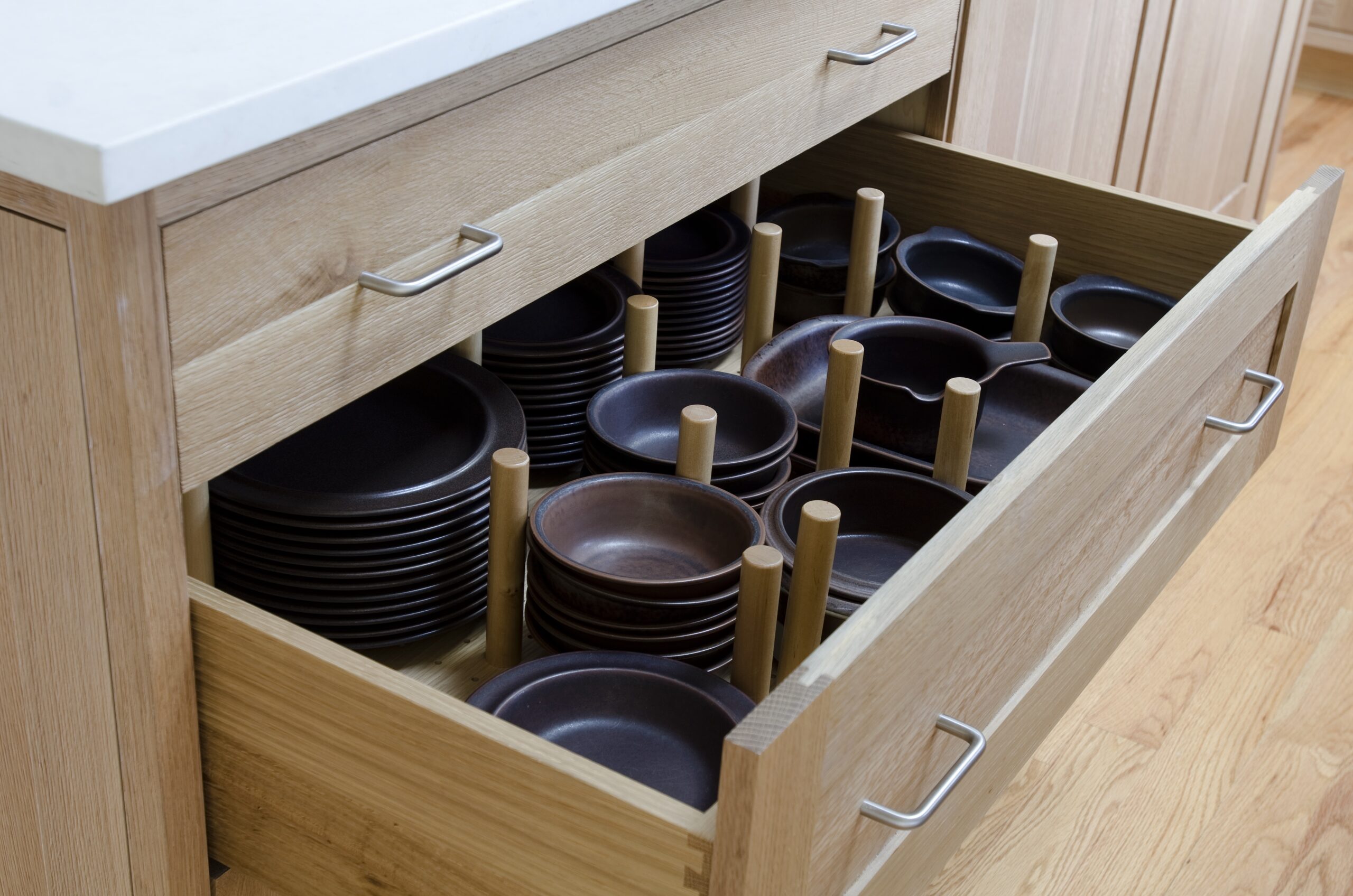 Open wooden kitchen drawer with neatly organized dark brown plates and bowls held in place by wooden pegs; light countertop and cabinets showcase the elegance of luxury interior design.