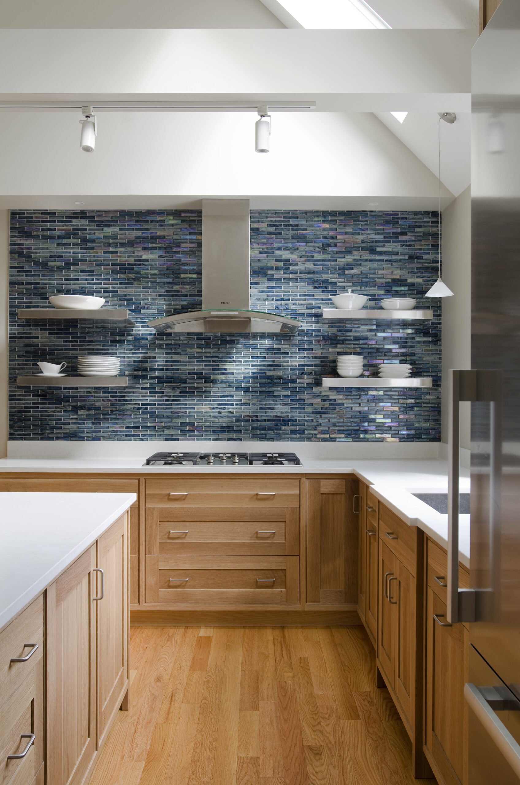 Modern kitchen with wood cabinets, a blue mosaic tile backsplash, stainless steel range hood, floating shelves with white dishes, and hardwood floors—crafted with a luxury interior design touch by an expert Interior Designer Massachusetts.