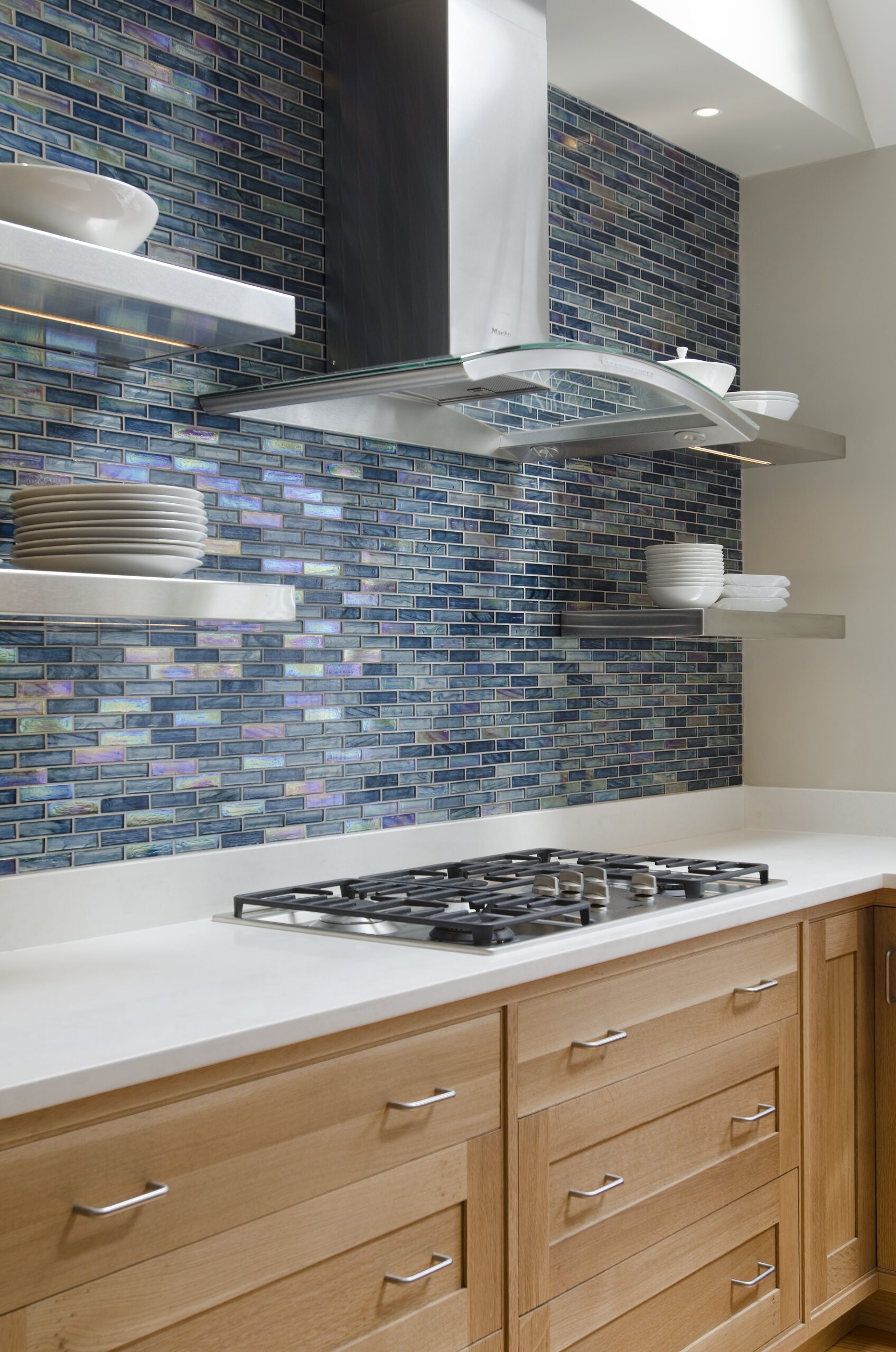 Modern kitchen by an Interior Designer in Massachusetts featuring a gas cooktop, wooden cabinets, blue glass tile backsplash, metal range hood, white countertop, and open shelves displaying white dishes—perfect for luxury interior design.