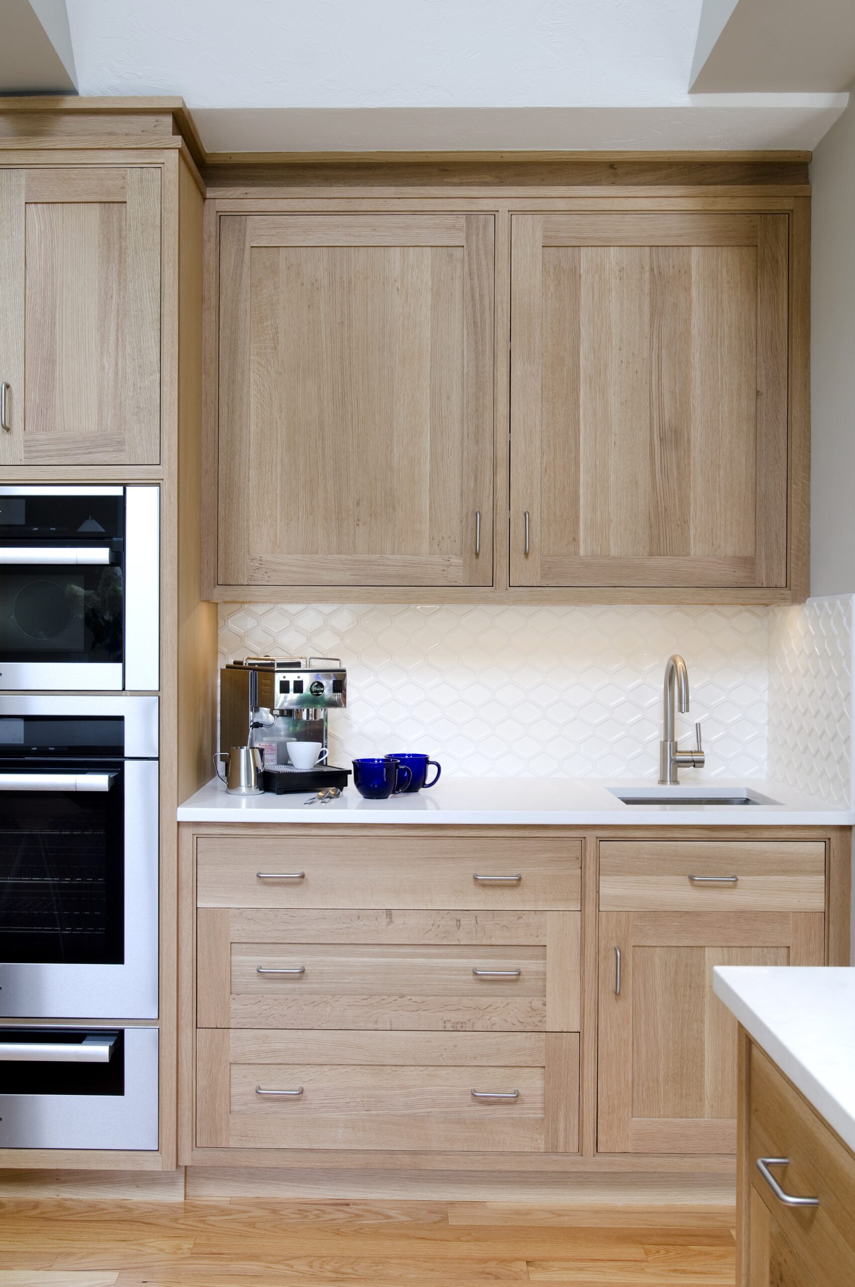 Modern kitchen corner with light wood cabinets, white countertop, small sink, built-in oven, and a coffee machine beside blue mugs against a white tile backsplash—perfect for those who appreciate luxury interior design.