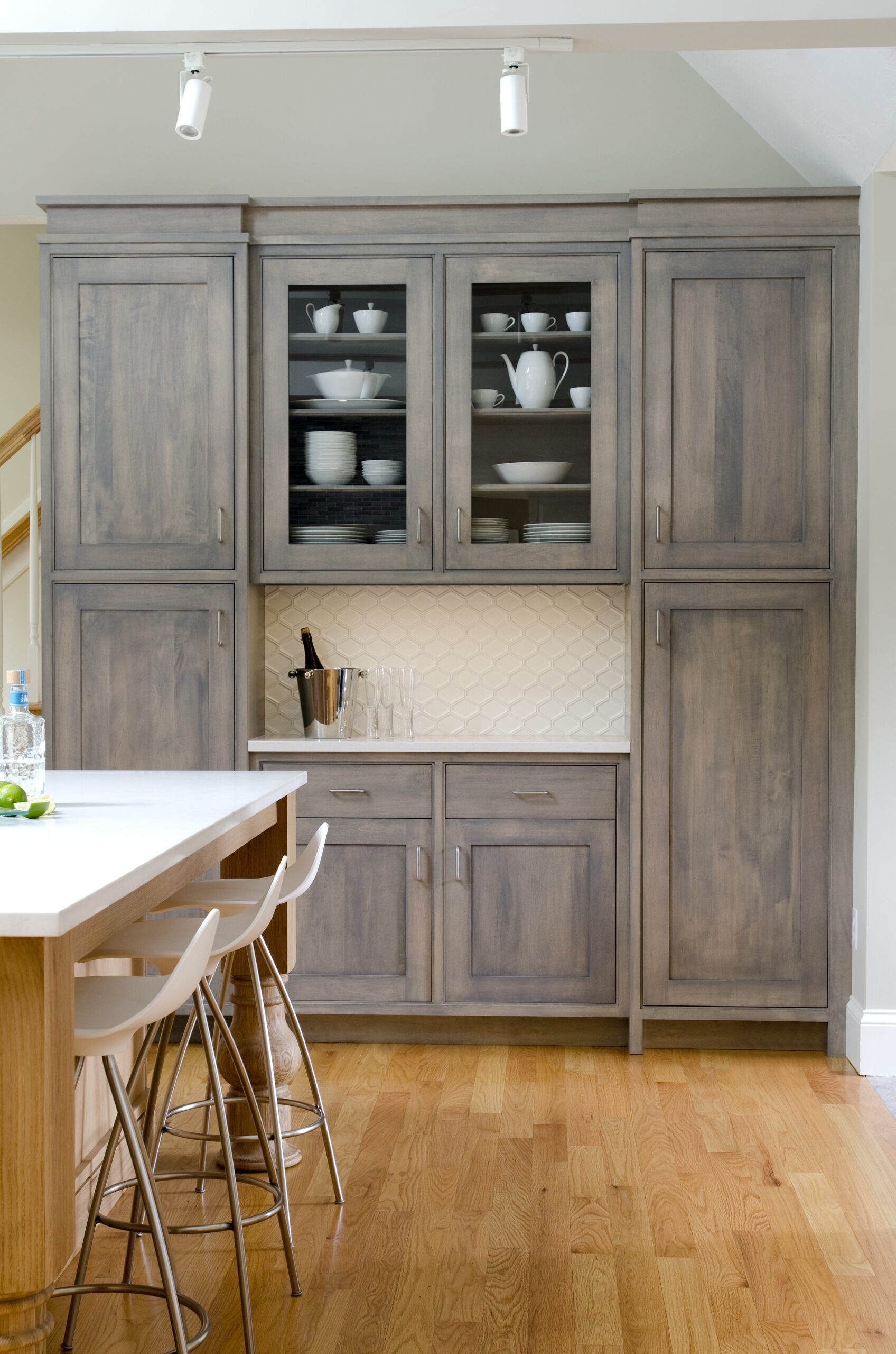 A kitchen with light wood cabinets, glass-front upper doors displaying white dishes, a tiled backsplash, and an island with white stools on a hardwood floor—perfectly curated for luxury interior design by an expert Interior Designer in Massachusetts.