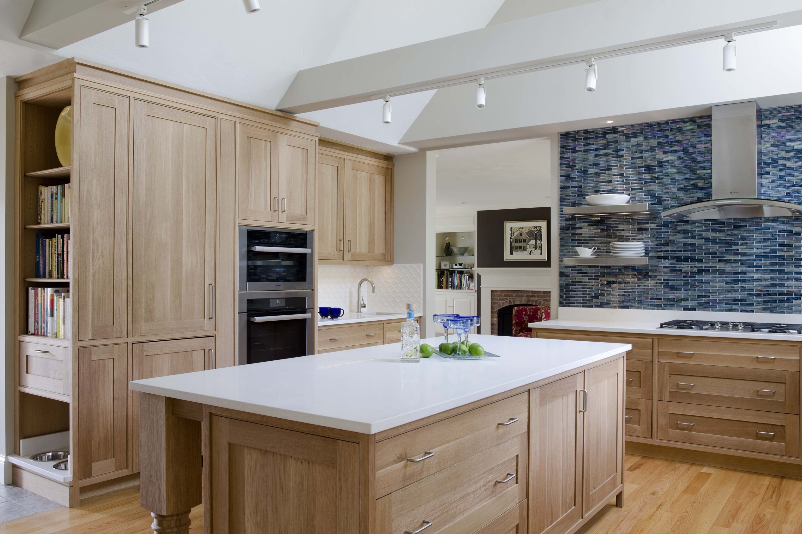 Modern kitchen with light wood cabinets, white countertops, a central island, blue mosaic tile backsplash, and stainless steel appliances under ceiling track lighting—showcasing luxury interior design by an expert Interior Designer in Massachusetts.