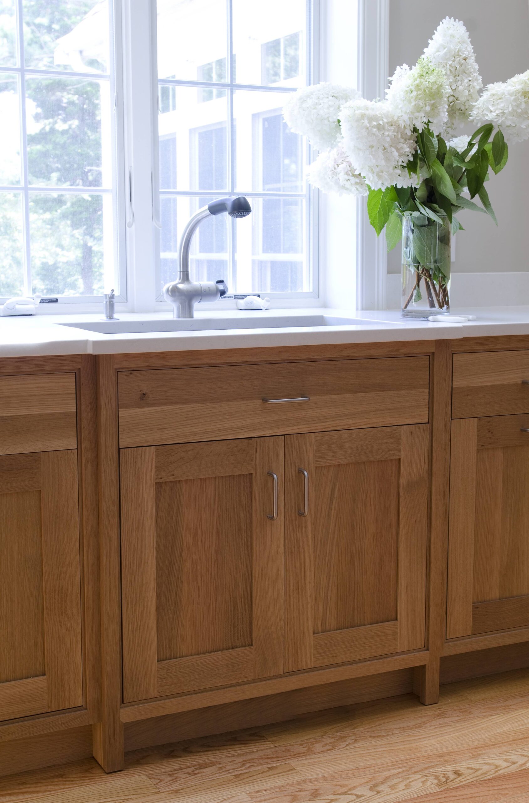 A kitchen sink with a modern faucet is set in a white countertop with wooden cabinets below. Styled by a luxury interior design expert, a glass vase with white flowers sits on the counter near a large window.