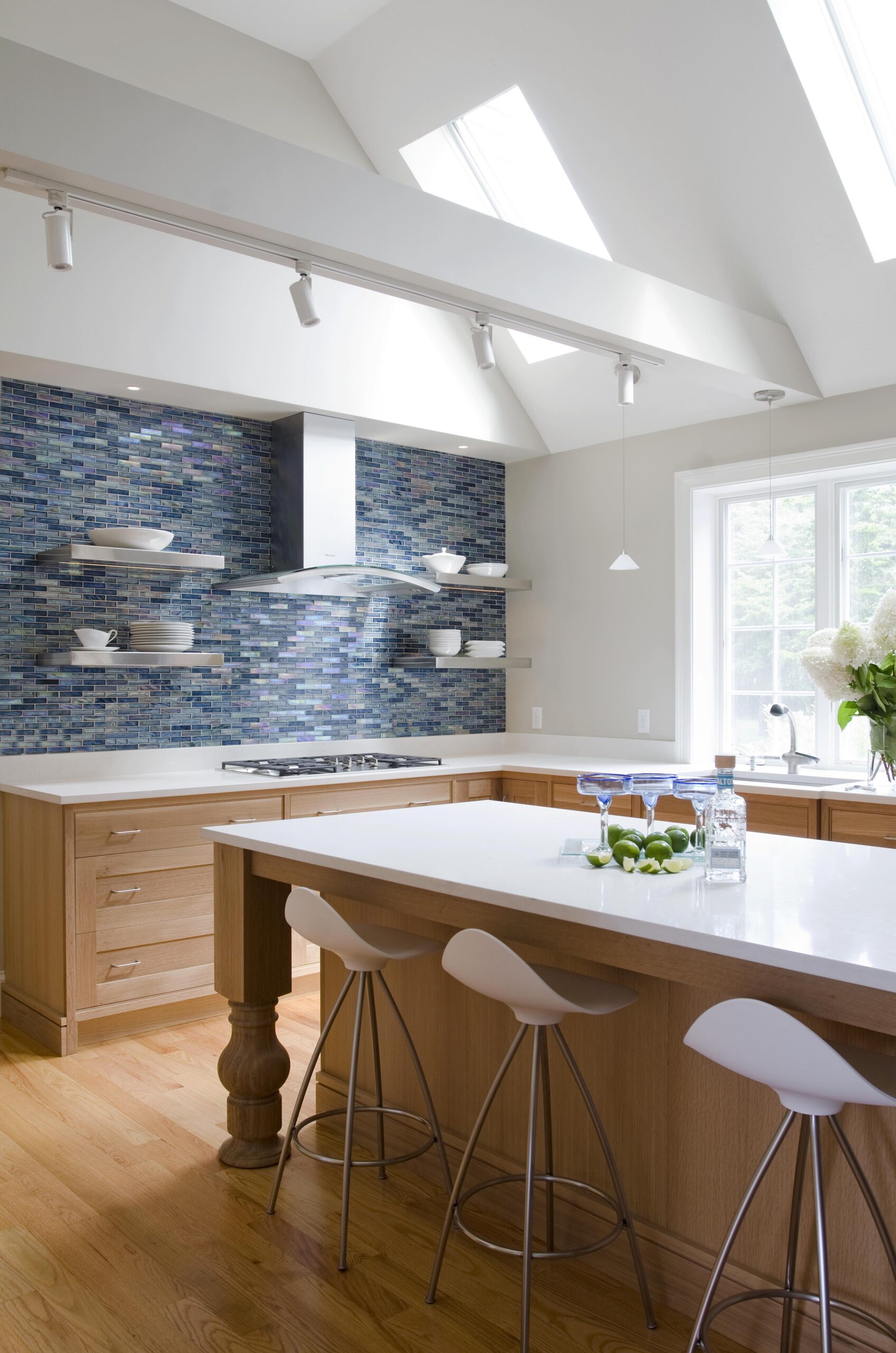 Modern kitchen with light wood cabinets, white countertops, blue mosaic tile backsplash, open shelves, bar stools, and natural light from skylights and a window—an example of Luxury Interior Design by an expert Interior Designer Massachusetts.