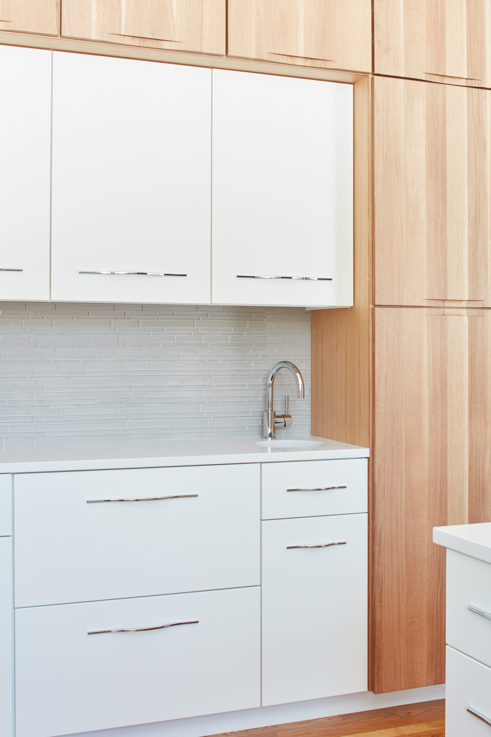 Modern kitchen with white cabinets, stainless steel handles, light wood paneling, a silver faucet, and a light gray tile backsplash—crafted for those seeking luxury interior design by a leading interior designer in Massachusetts.