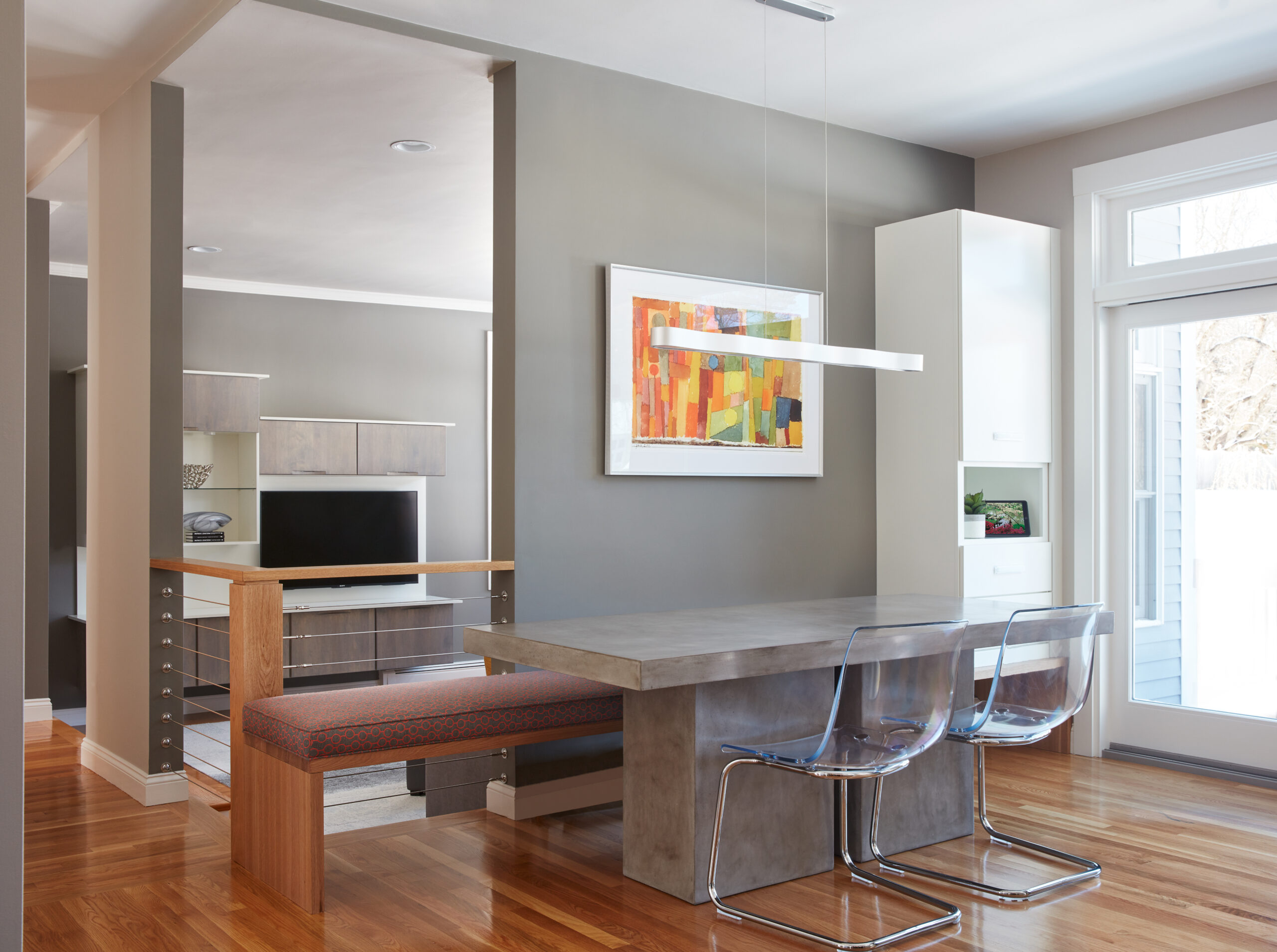 Modern dining area with a concrete table, two clear plastic chairs, a built-in bench, abstract wall art, and large windows letting in natural light—showcasing the elegance of luxury interior design.