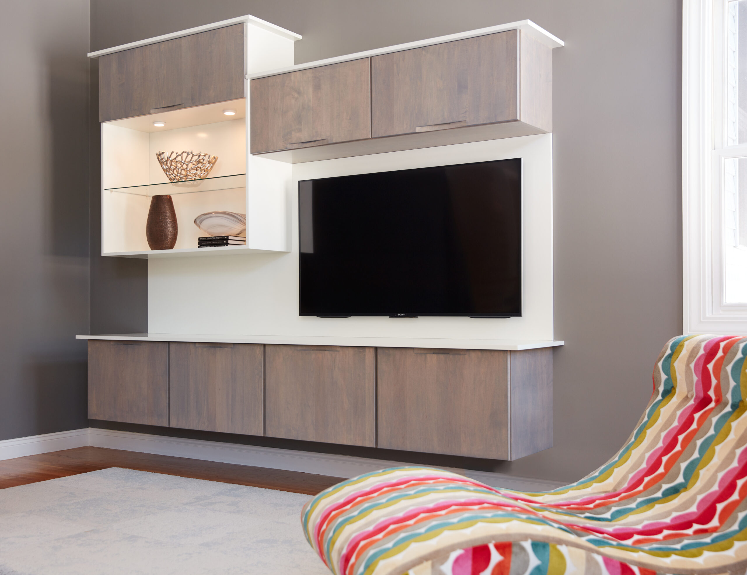 A modern living room inspired by luxury interior design features a wall-mounted TV, wooden storage cabinets, display shelves with decor items, a striped lounge chair, and a window letting in natural light.