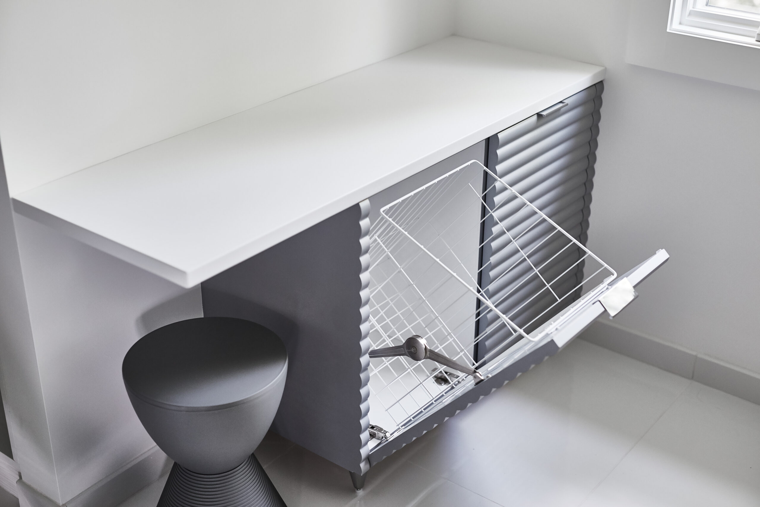 A laundry hamper with a pull-out wire basket is partially open below a white countertop in a modern room with light flooring and a small grey stool, showcasing touches of luxury interior design.