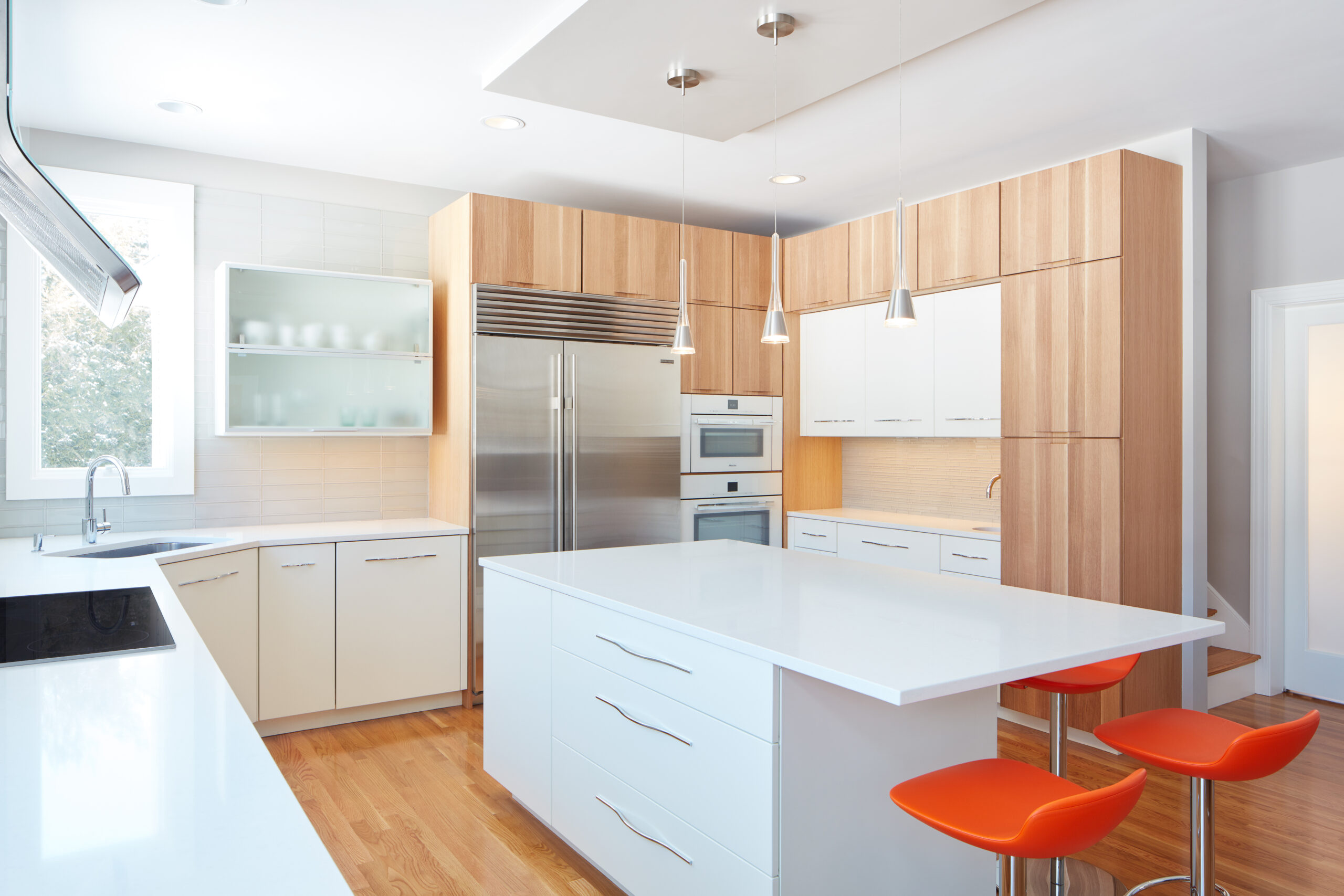 Modern kitchen with white countertops, wood cabinetry, stainless steel appliances, an island with drawers, and three orange barstools—showcasing the touch of a luxury interior design expert or interior designer Massachusetts.