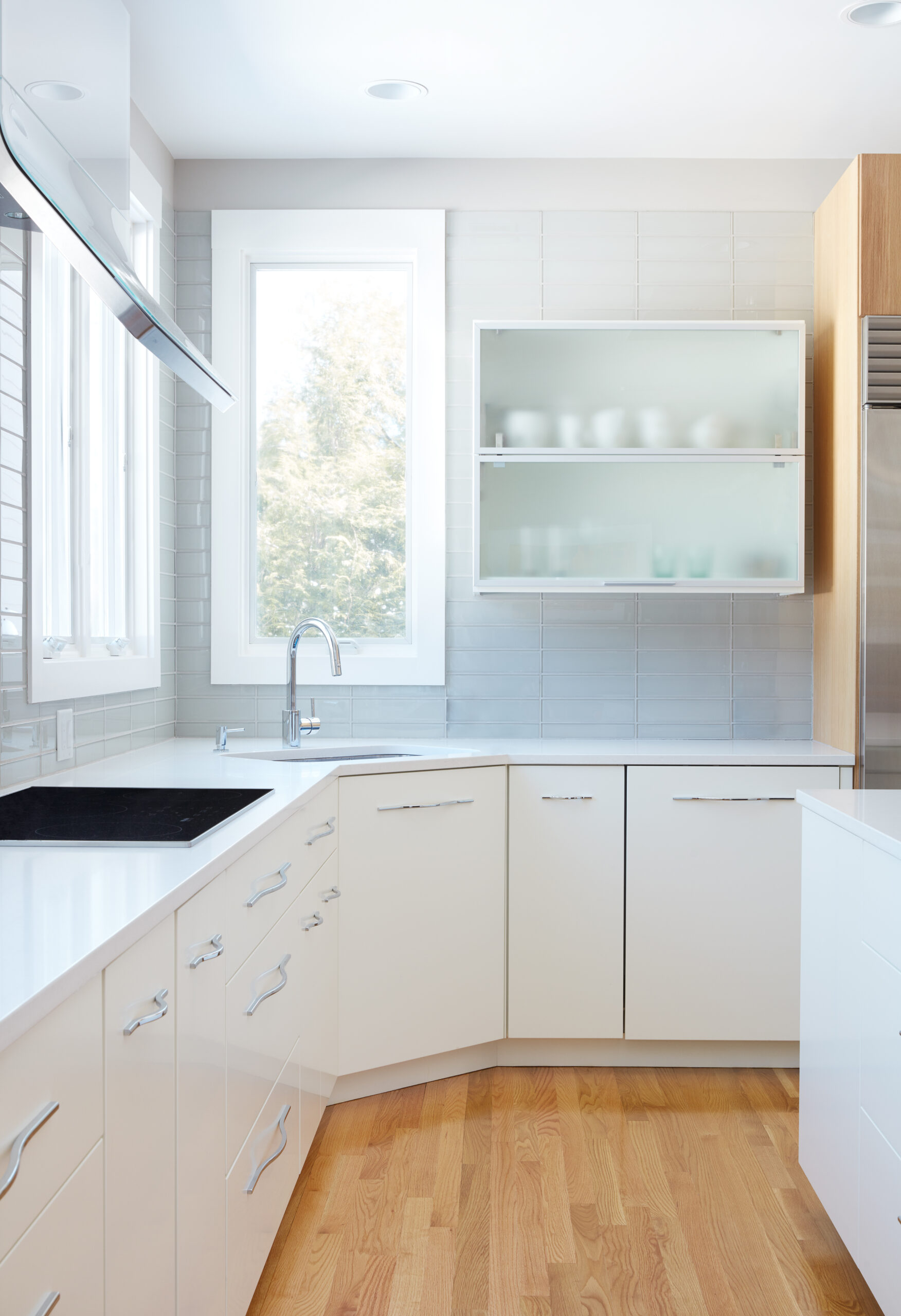 Modern kitchen with white cabinets, frosted glass upper cabinet, stainless steel appliances, large windows, and light wood flooring—all elevated by a touch of luxury interior design. Natural light fills the space for a bright, inviting atmosphere.