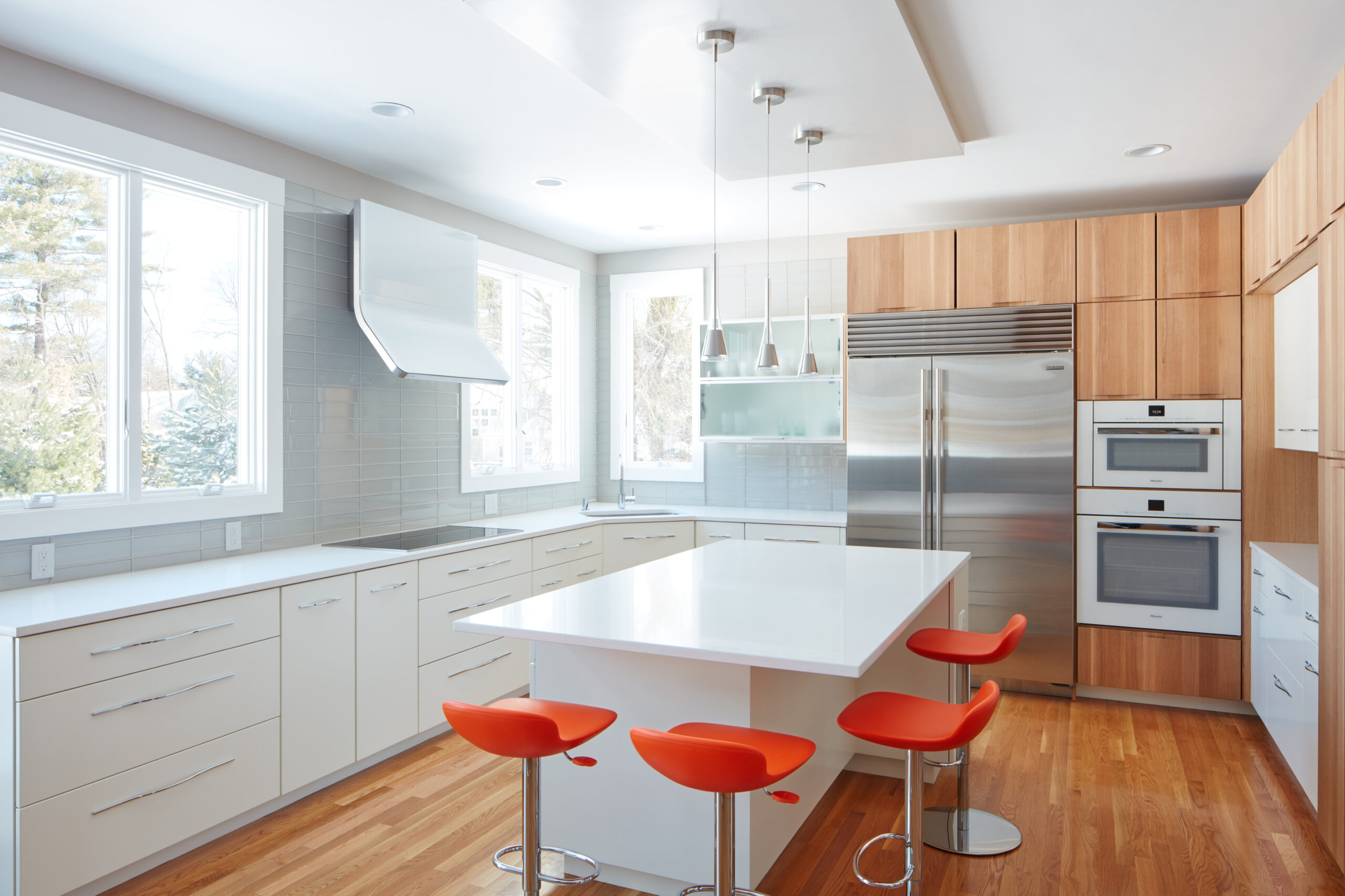 Modern kitchen with white cabinets, stainless steel appliances, a large island, orange bar stools, and hardwood floors. Large windows provide natural light—a stunning example of luxury interior design by a top interior designer in Massachusetts.