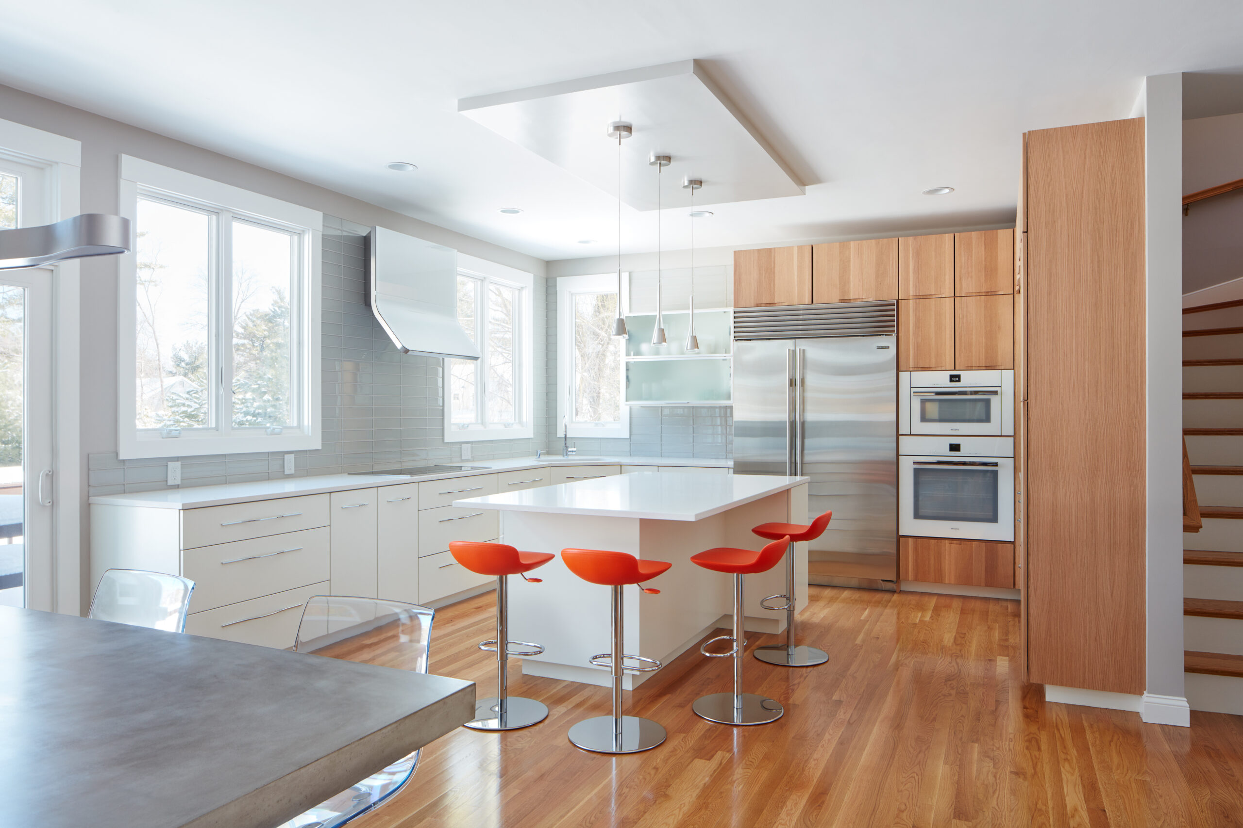 Modern kitchen with white cabinets, stainless steel appliances, an island with four orange bar stools, wood flooring, and large windows—a stunning example of luxury interior design by a top interior designer in Massachusetts.