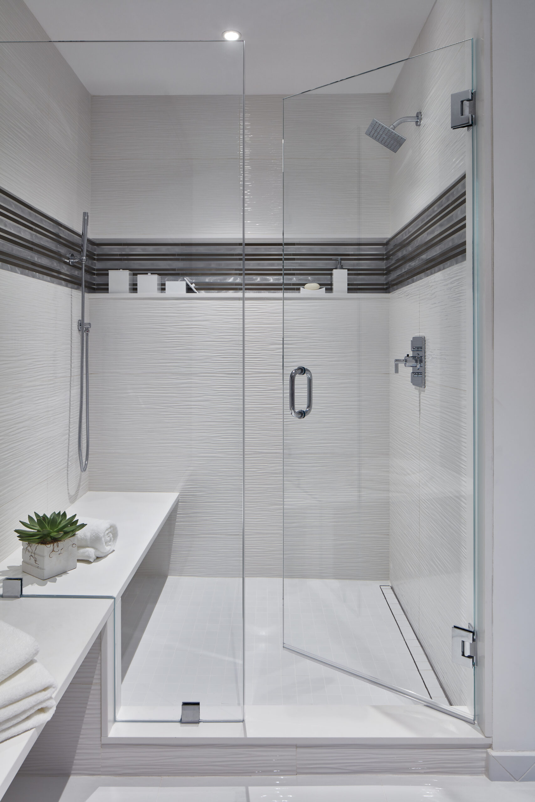 Modern walk-in shower with glass door, white tiles, a built-in bench, gray horizontal accent stripes, rain showerhead, and minimalistic toiletries—showcasing luxury interior design for a refined touch by an expert interior designer in Massachusetts.