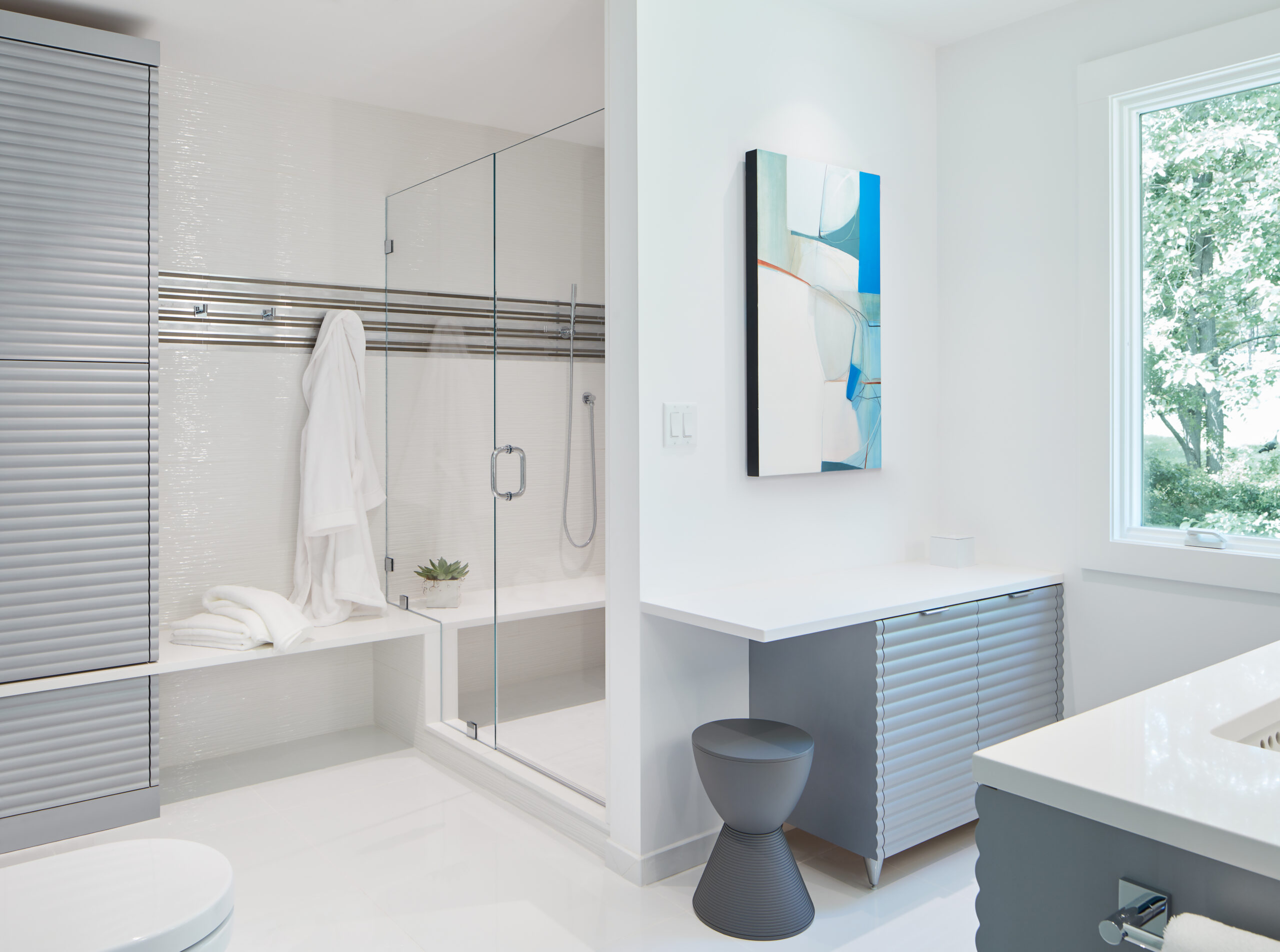 Experience luxury interior design in this modern bathroom featuring a glass-enclosed shower, sleek cabinetry, abstract wall art, and a large window with lush views—expertly crafted by an interior designer Massachusetts residents trust.