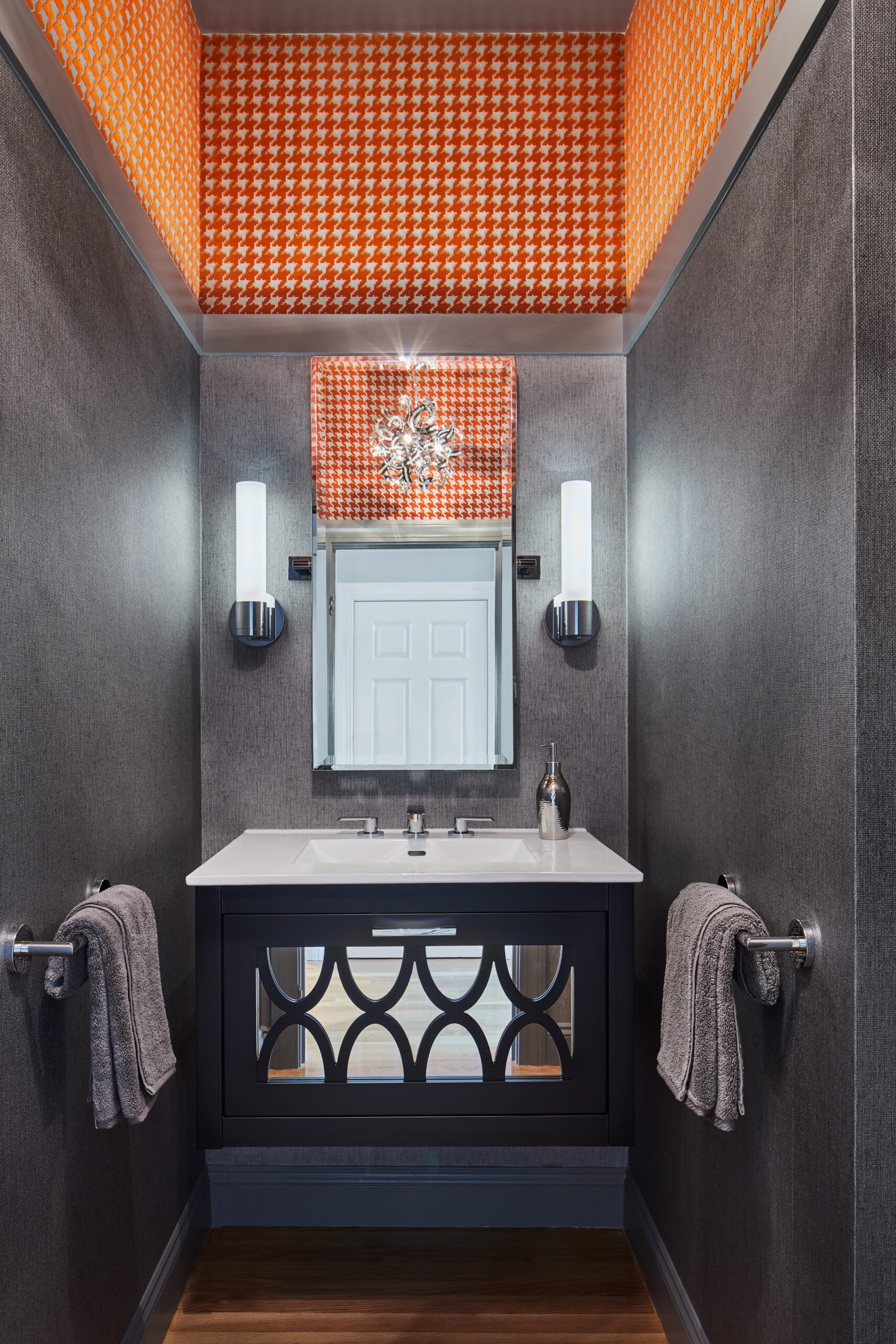 Modern bathroom with a dark vanity, two gray hand towels, wall-mounted lights, a rectangular mirror, and orange houndstooth-patterned wallpaper—showcasing luxury interior design by an expert interior designer in Massachusetts.