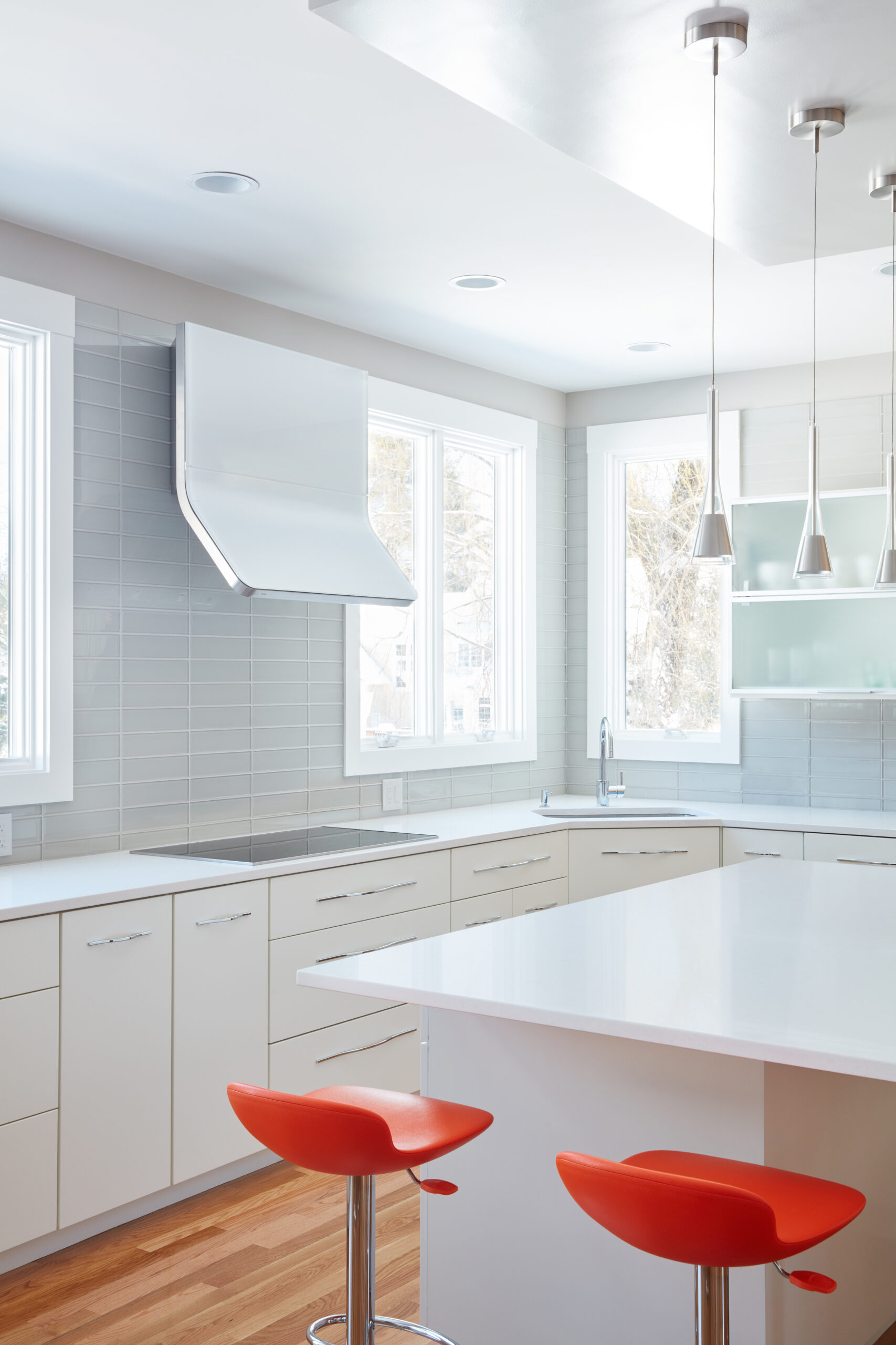 Modern kitchen with white cabinets, gray tile backsplash, white countertops, pendant lights, and two red bar stools at the island; large windows provide natural light—perfect for luxury interior design by an experienced interior designer in Massachusetts.