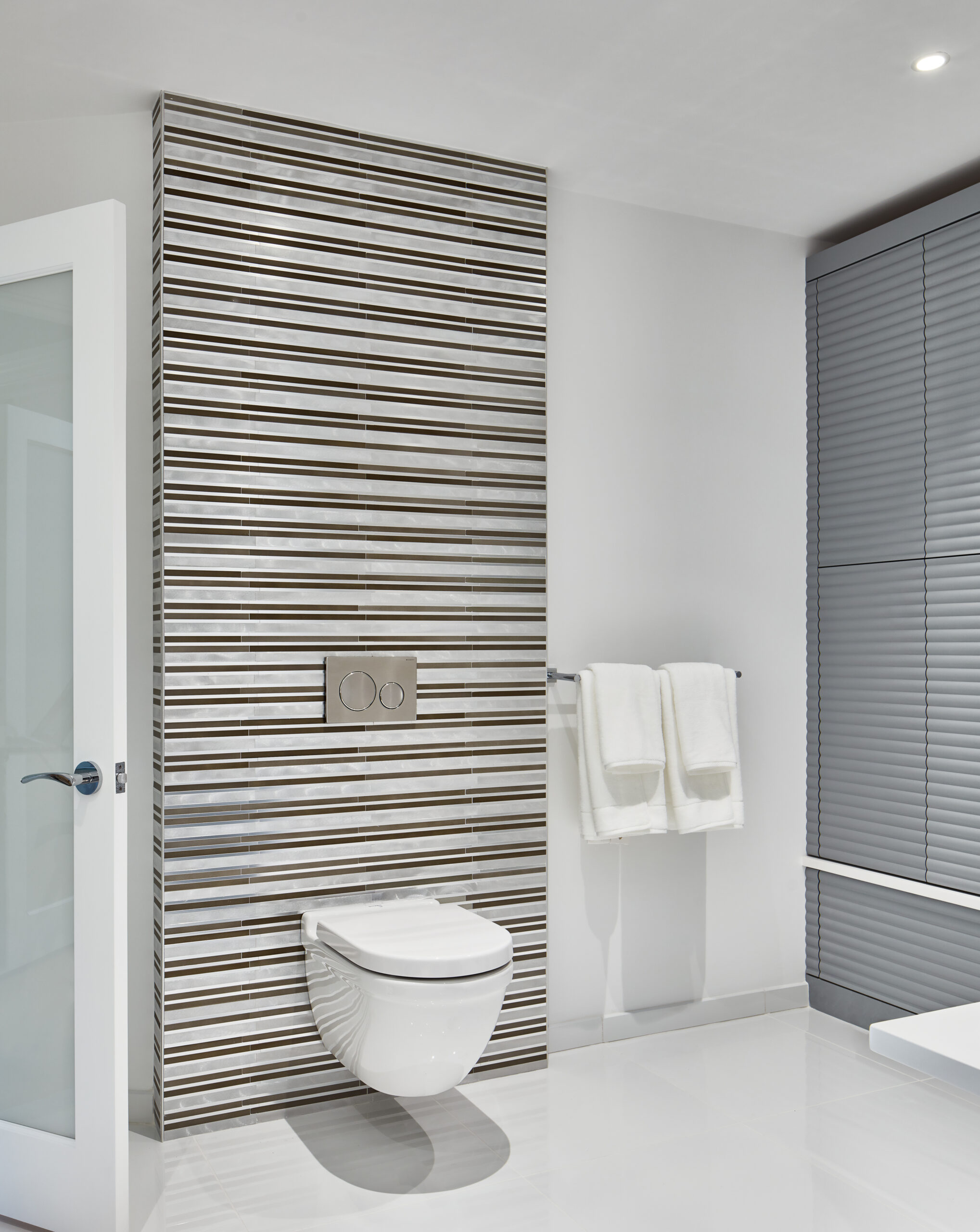 Modern bathroom with a wall-mounted toilet, striped tile accent wall, dual flush buttons, and two white towels on a rack—perfectly showcasing luxury interior design by an expert interior designer in Massachusetts.