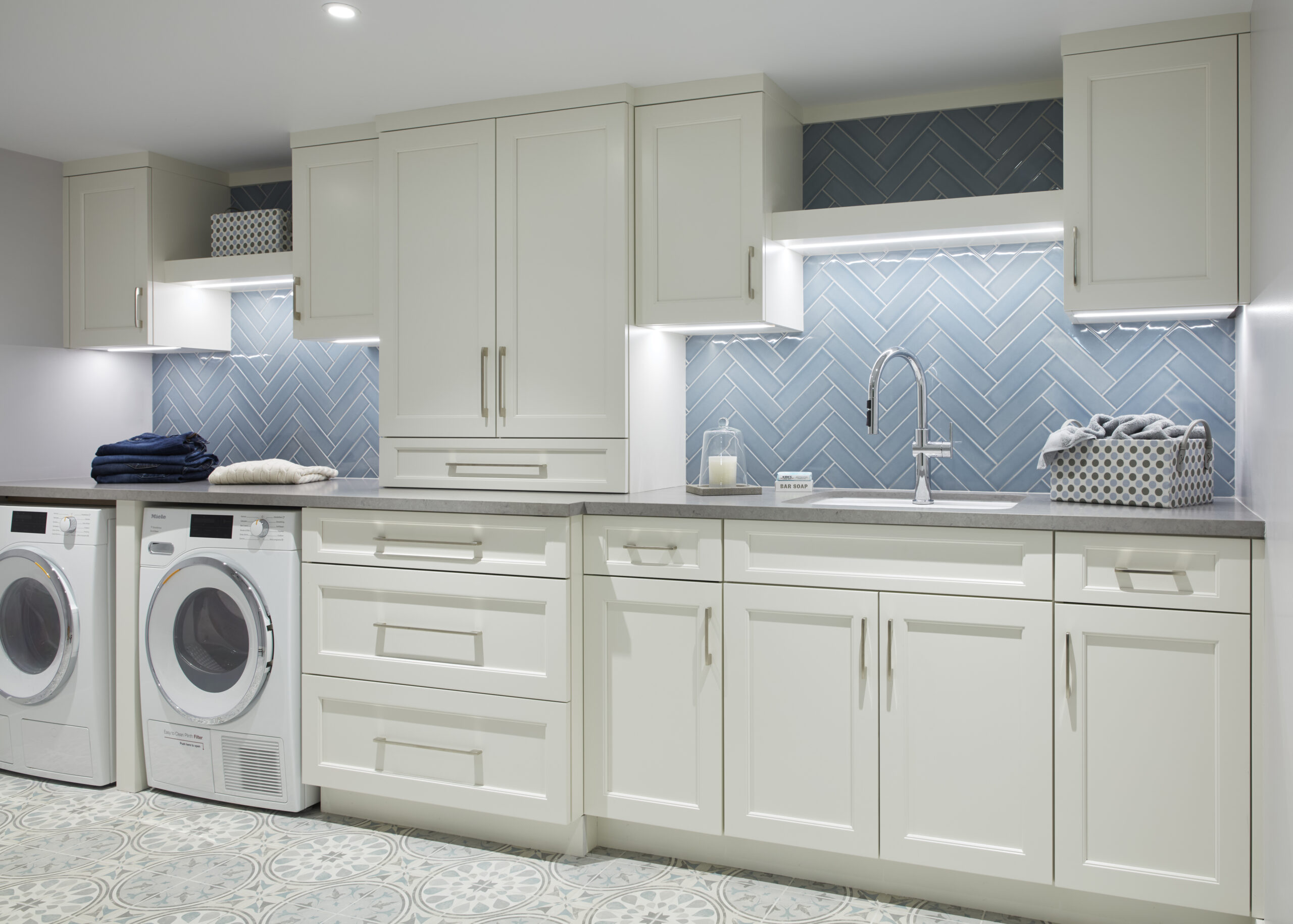 Modern laundry room with white cabinets, a front-loading washer and dryer, gray countertop, blue herringbone tile backsplash, and patterned floor tiles—crafted with a touch of luxury interior design by an expert interior designer in Massachusetts.