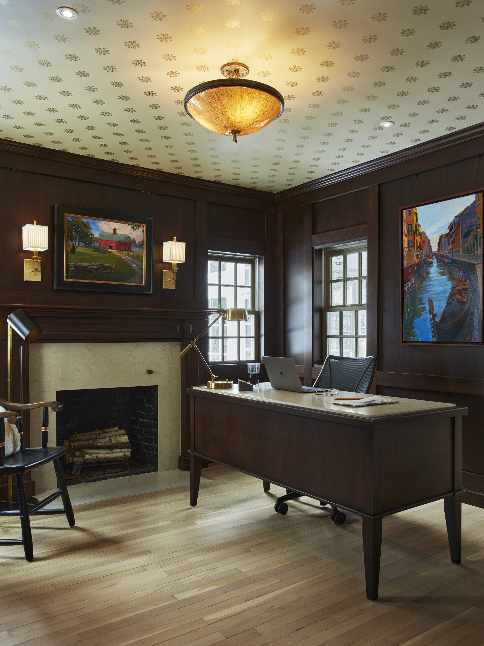 A traditional office with dark wood paneling, a fireplace, a wooden desk with a laptop, and two colorful paintings on the walls showcases classic luxury interior design.