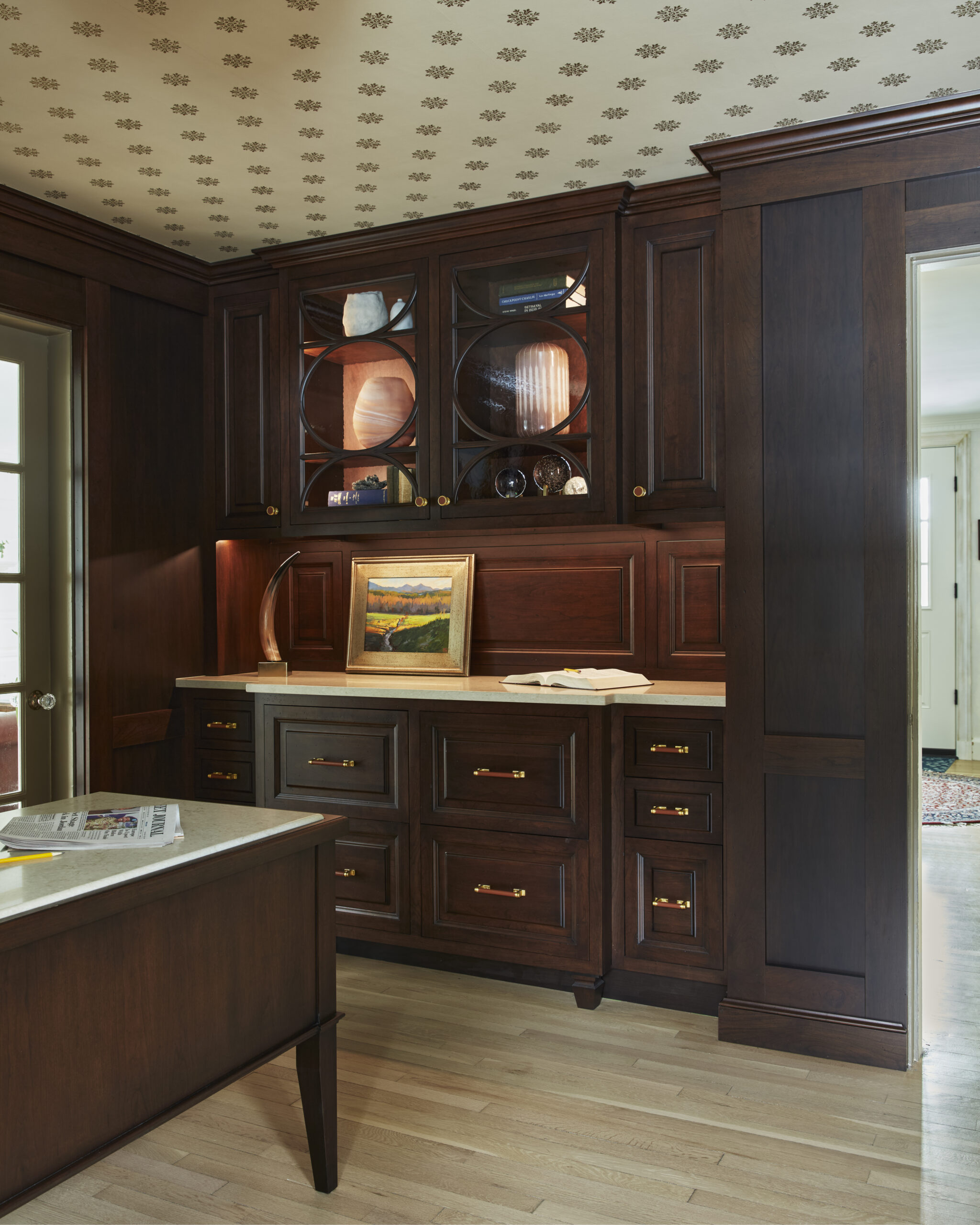 A kitchen with dark wood cabinets, glass-front upper cupboards, brass handles, a light wood floor, and a framed painting on the countertop under soft lighting exudes the elegance of luxury interior design.