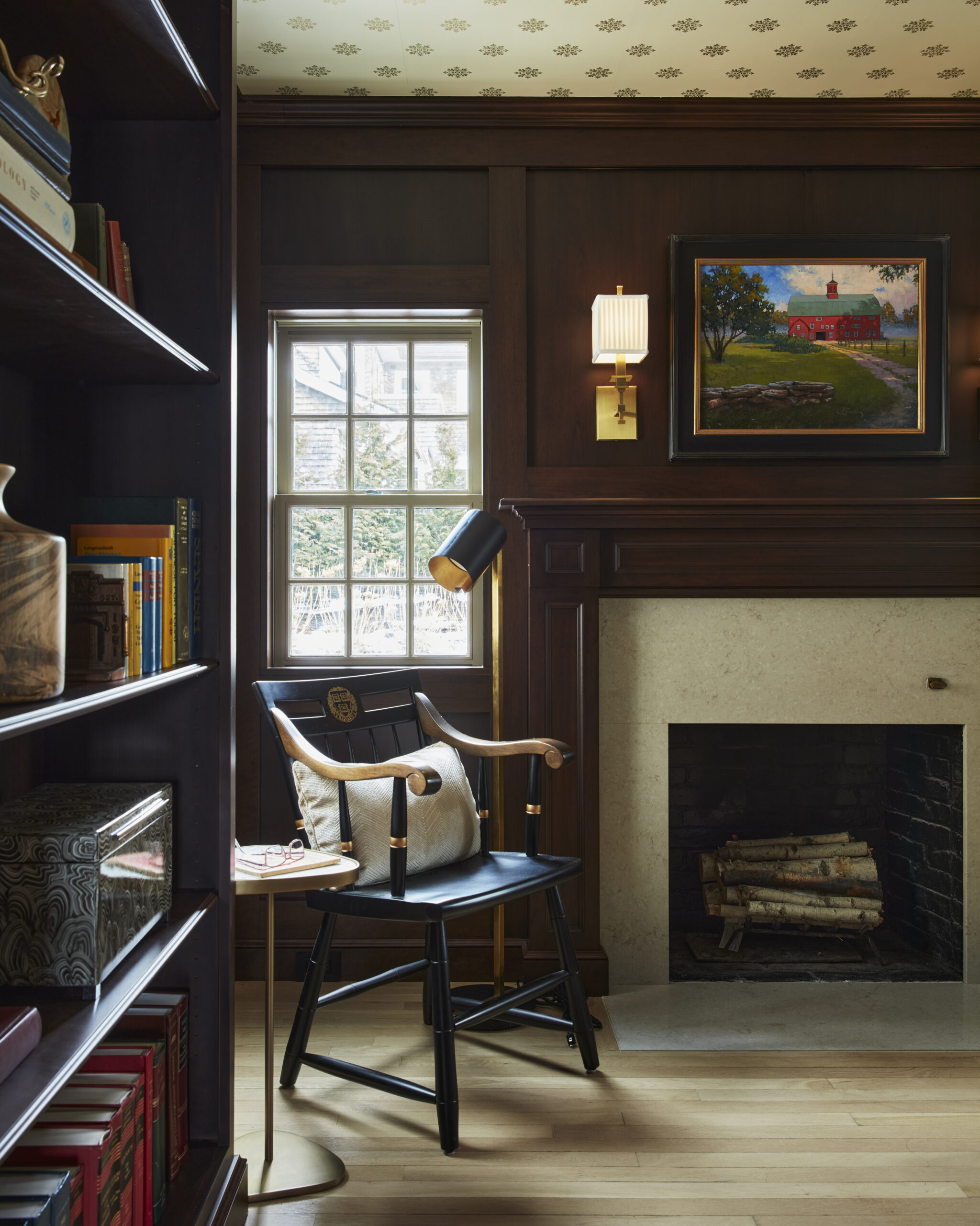 A reading corner in a wood-paneled room with a black chair, side table, fireplace with logs, bookshelf, window, and framed painting above the mantel exudes Luxury Interior Design for a truly refined atmosphere.