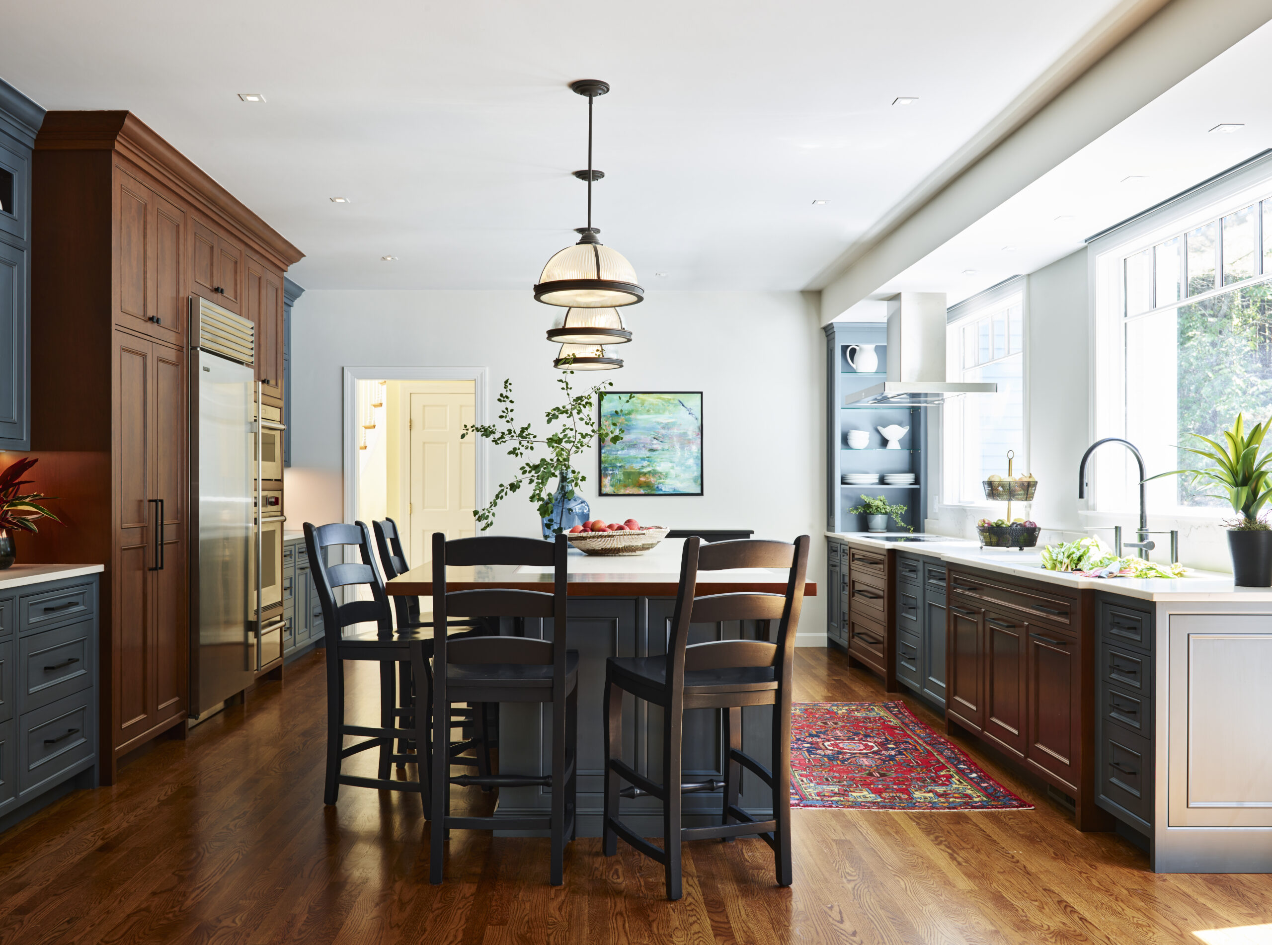 Spacious kitchen with dark wood cabinets, island with chairs, pendant lights, large windows, stainless steel appliances, and decorative plants—showcasing luxury interior design by a top interior designer in Massachusetts.
