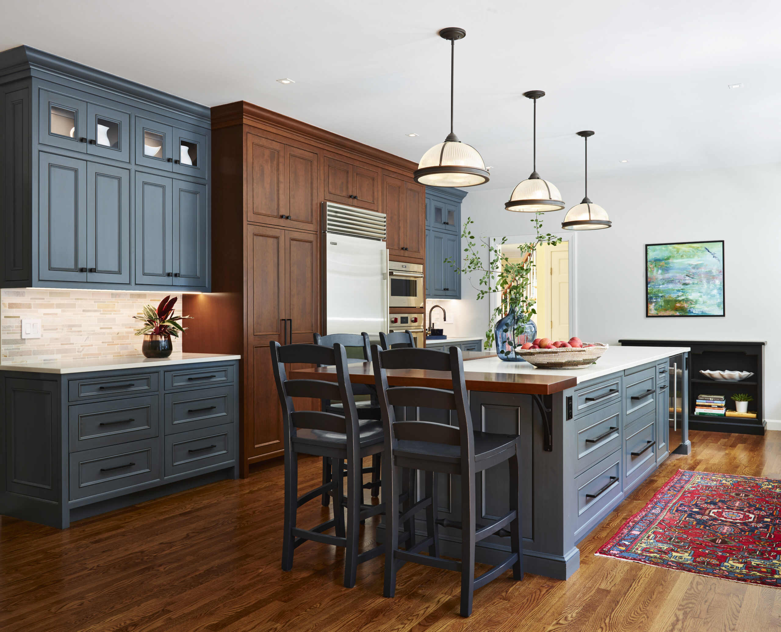 Modern kitchen with blue and wood cabinetry, stainless steel appliances, a large island with chairs, pendant lights, hardwood floors, and a colorful rug—showcasing luxury interior design by an expert interior designer in Massachusetts.