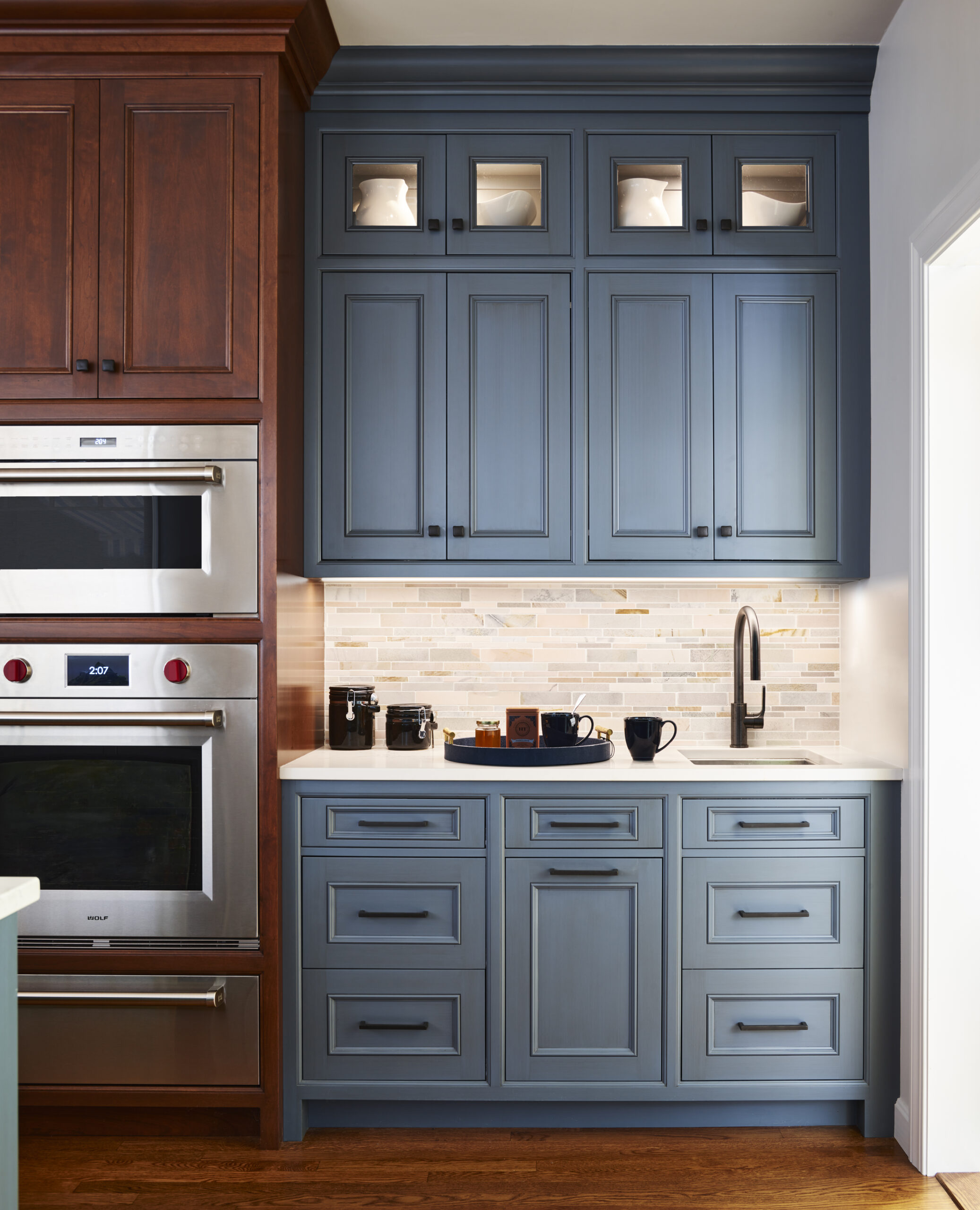 A kitchen by an Interior Designer Massachusetts features blue cabinets, a white countertop, a modern faucet, tile backsplash, and built-in stainless steel ovens beside brown cabinetry for a touch of luxury interior design.