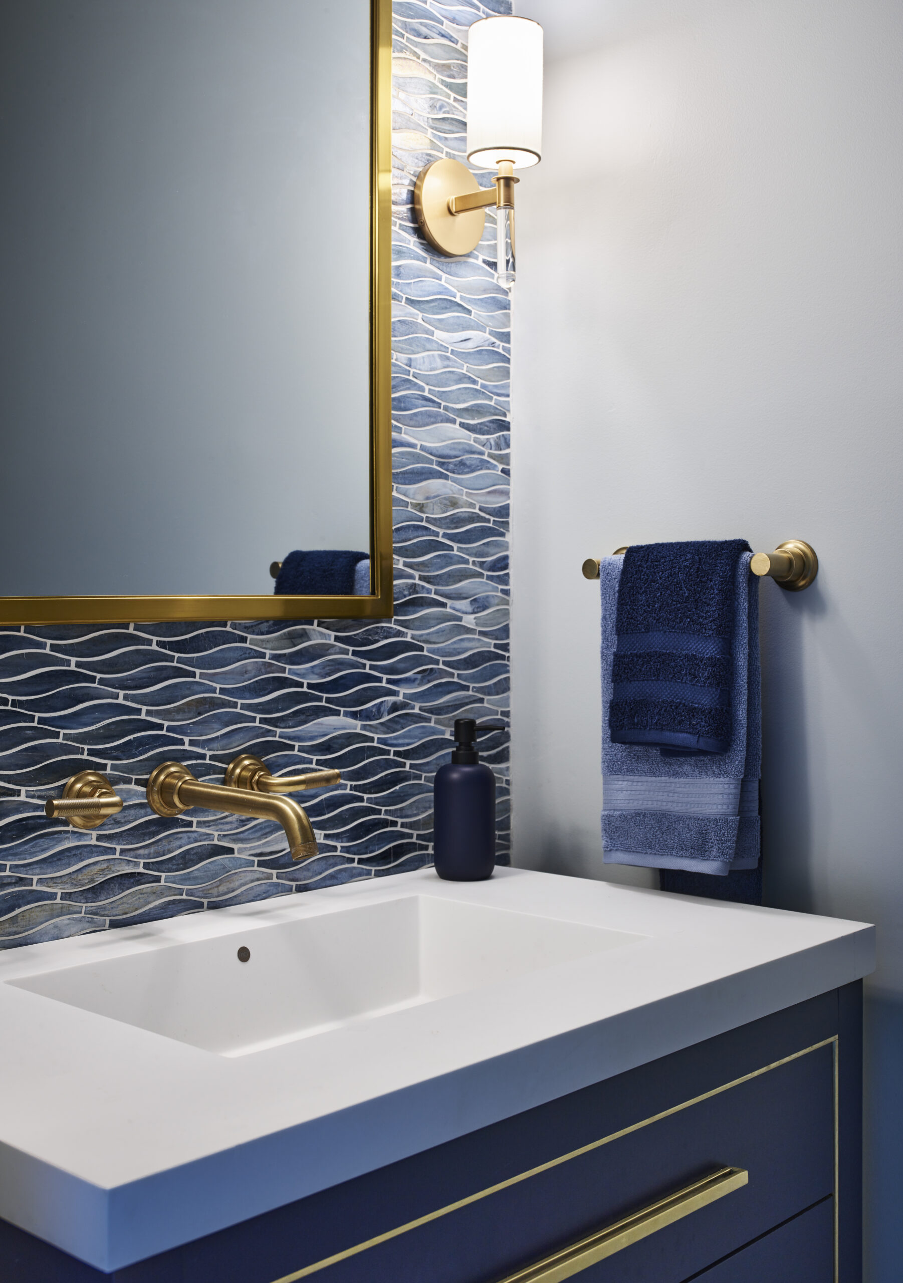 Modern bathroom with a rectangular mirror, brass fixtures, blue patterned tile backsplash, wall sconce, and blue vanity showcases Luxury Interior Design. Neatly folded navy towels rest on a gold towel bar for an elegant finish.