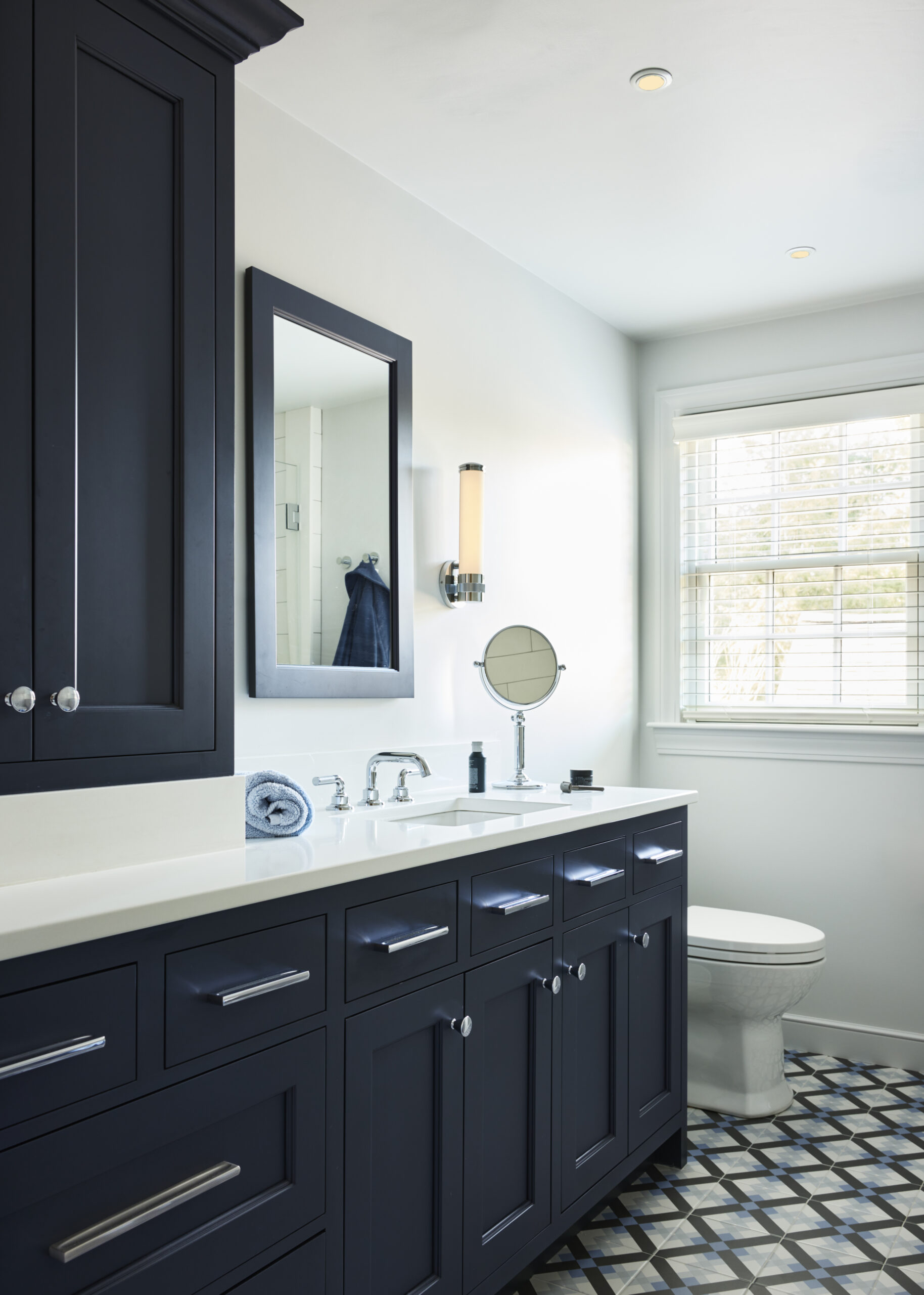 Modern bathroom with dark cabinets, a large mirror, a sink with a rolled towel, a round vanity mirror, and patterned floor tiles. Natural light enters through window blinds—an example of Luxury Interior Design by an Interior Designer Massachusetts.