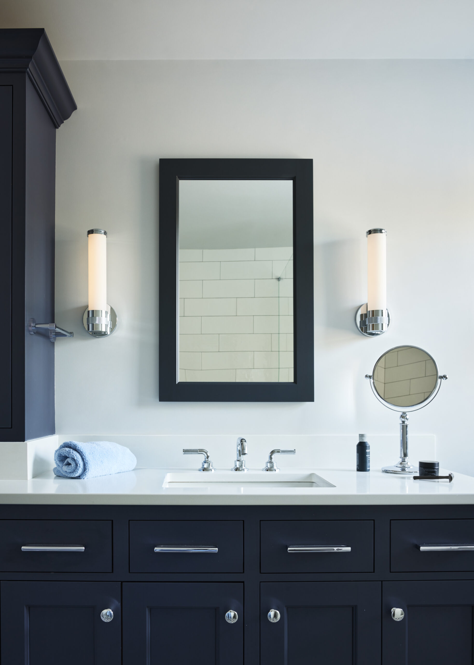 Modern bathroom vanity with dark cabinets, a rectangular mirror, two wall-mounted lights, a rolled blue towel, a small round mirror, and toiletries on a white countertop—perfect for those seeking luxury interior design.