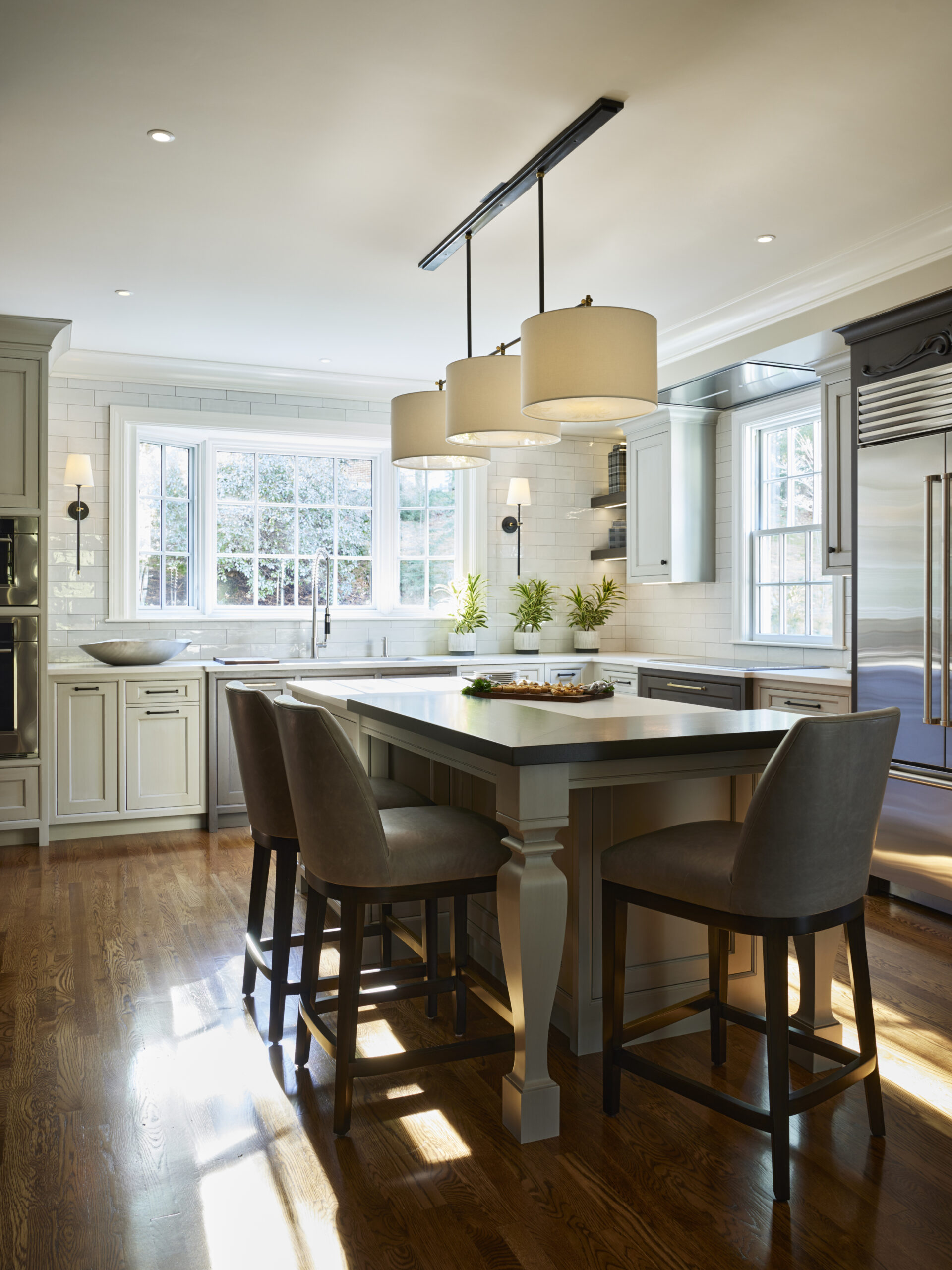 Bright kitchen with an island, three upholstered chairs, pendant lights, stainless steel appliances, and white cabinets—this Luxury Interior Design features large windows and hardwood floors by a top Interior Designer Massachusetts.