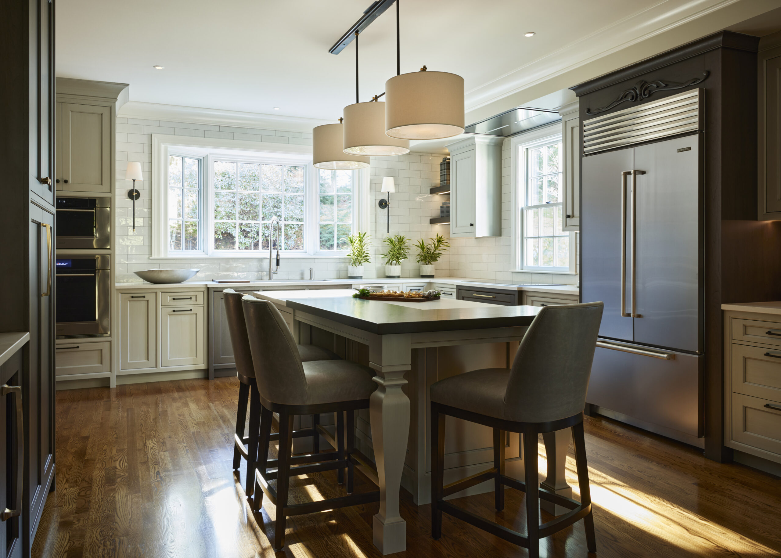 This luxury interior design features a modern kitchen with a central island, three pendant lights, stainless steel appliances, large windows, and a wooden floor. Two upholstered stools complete the look curated by an expert interior designer in Massachusetts.