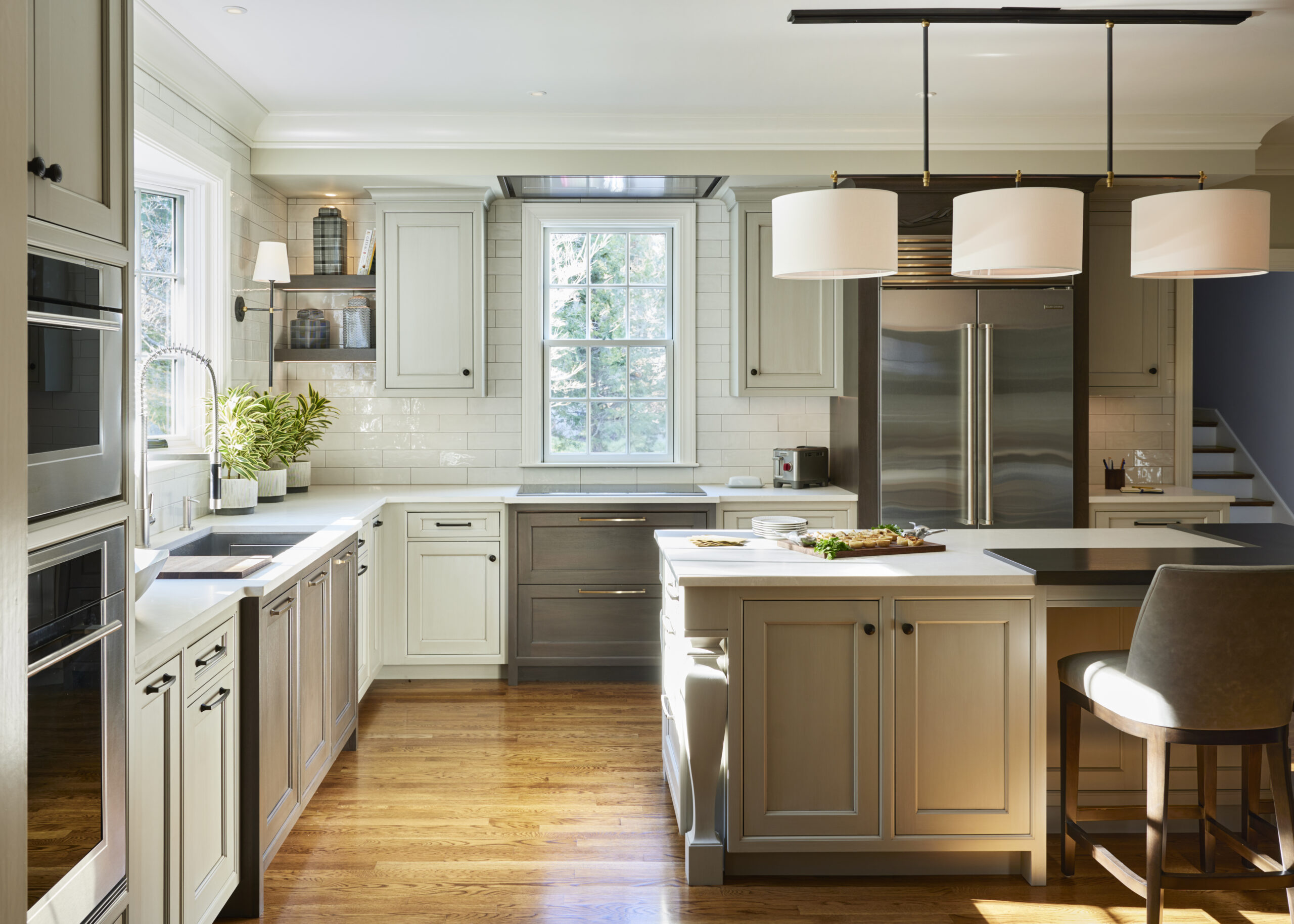 Modern kitchen with white cabinets, stainless steel appliances, a central island with seating, wood flooring, and sunlight streaming through a window—showcasing luxury interior design by an expert interior designer in Massachusetts.