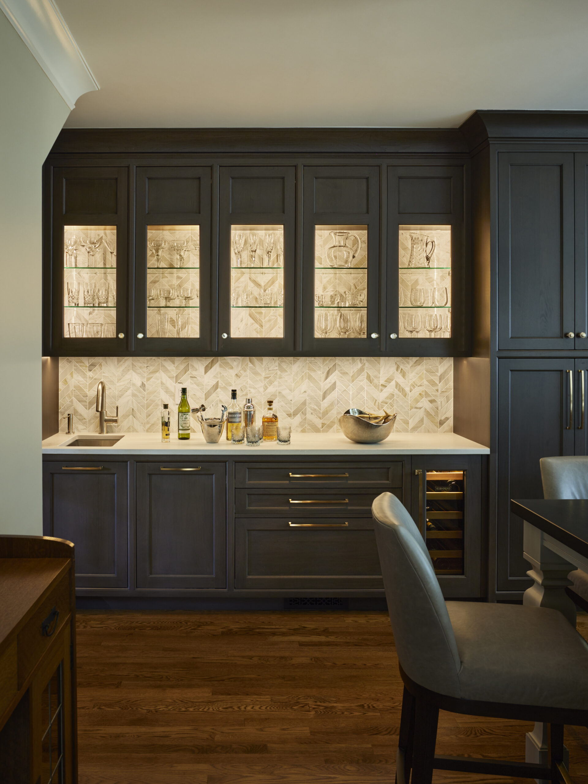 A home bar area with dark wood cabinets, glass-front uppers displaying glassware, a countertop with bottles, a sink, and a chevron-patterned backsplash—perfectly crafted for those who appreciate luxury interior design.