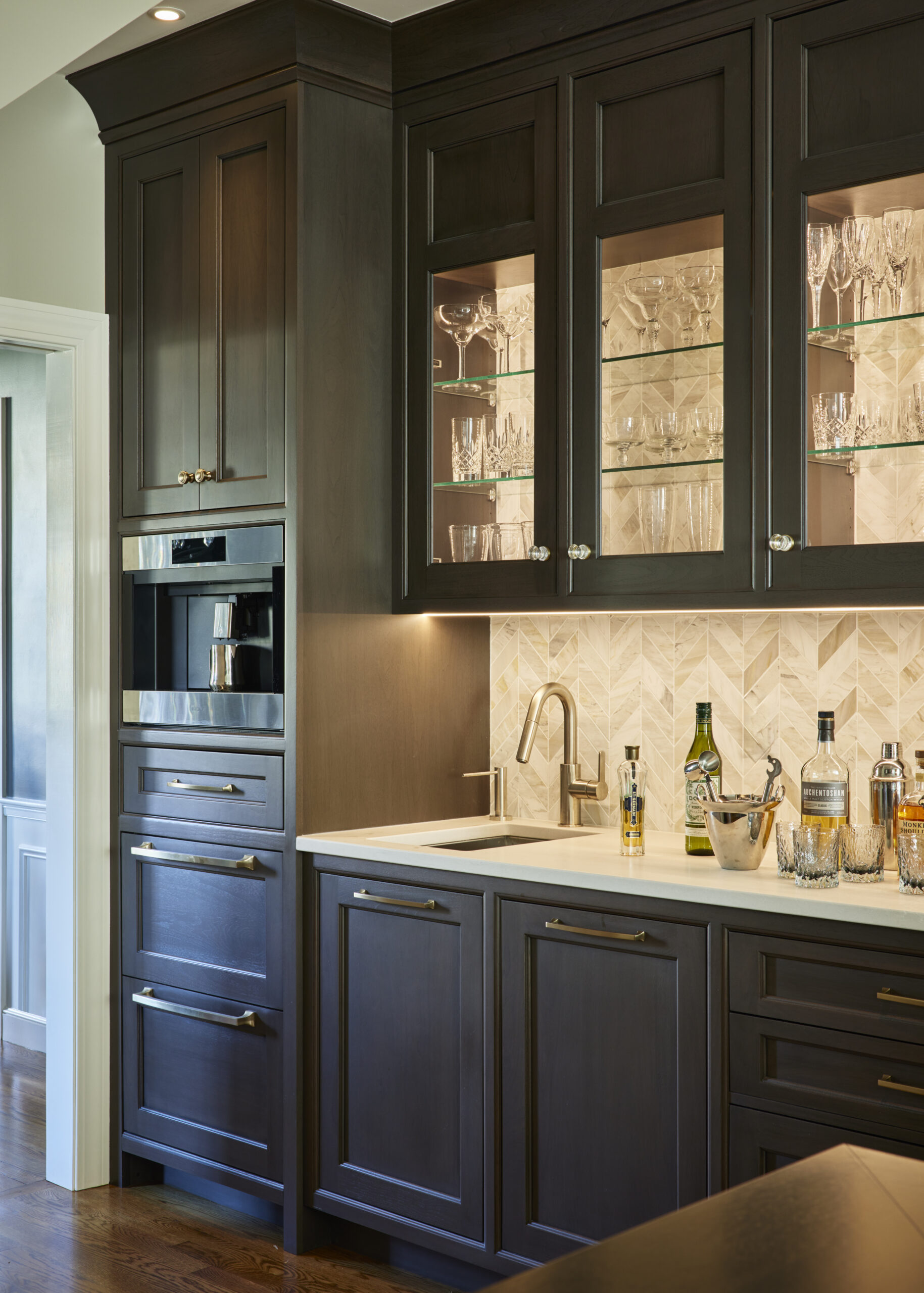 Modern kitchen bar area with dark wood cabinets, glass-front upper cupboards displaying glassware, a built-in oven, and a white countertop—an example of Luxury Interior Design by an expert Interior Designer Massachusetts.