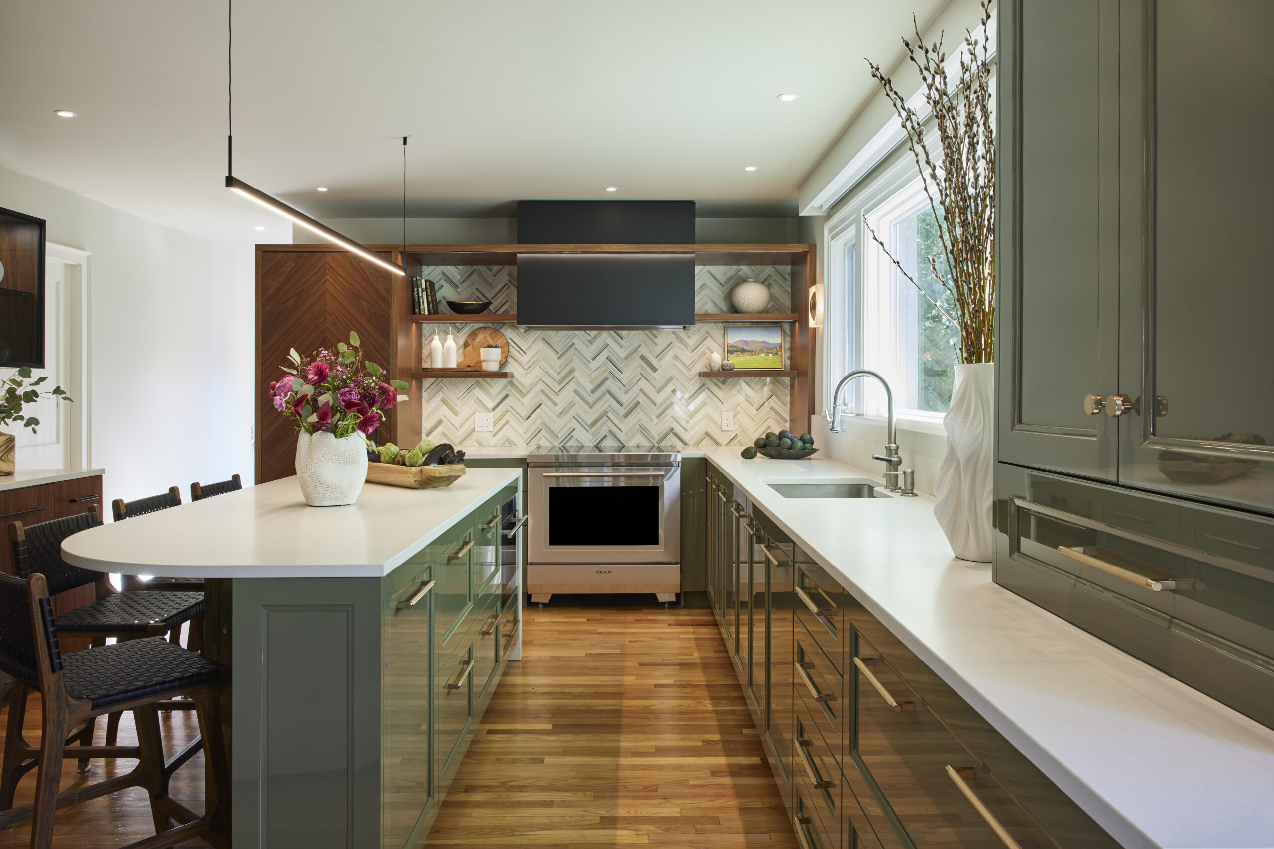 Modern kitchen with green cabinets, white countertops, a large island, built-in oven, patterned backsplash, wooden floor, and vases with flowers and branches—perfect for a Luxury Interior Design by an expert Interior Designer in Massachusetts.