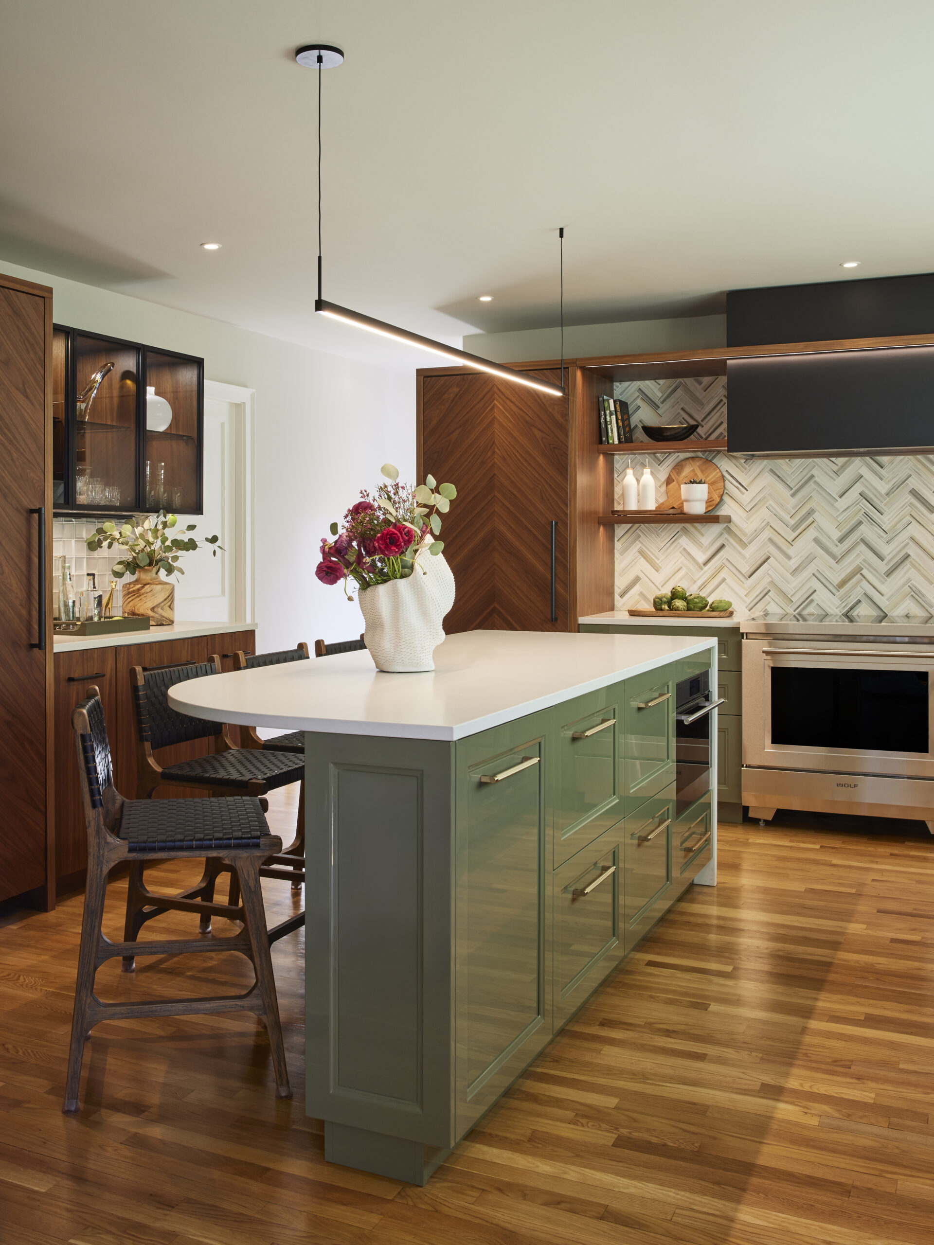 Modern kitchen with a green island, white countertop, wooden flooring, wood cabinets, herringbone tile backsplash, and a vase of flowers—showcasing the elegance of Luxury Interior Design by an Interior Designer Massachusetts.