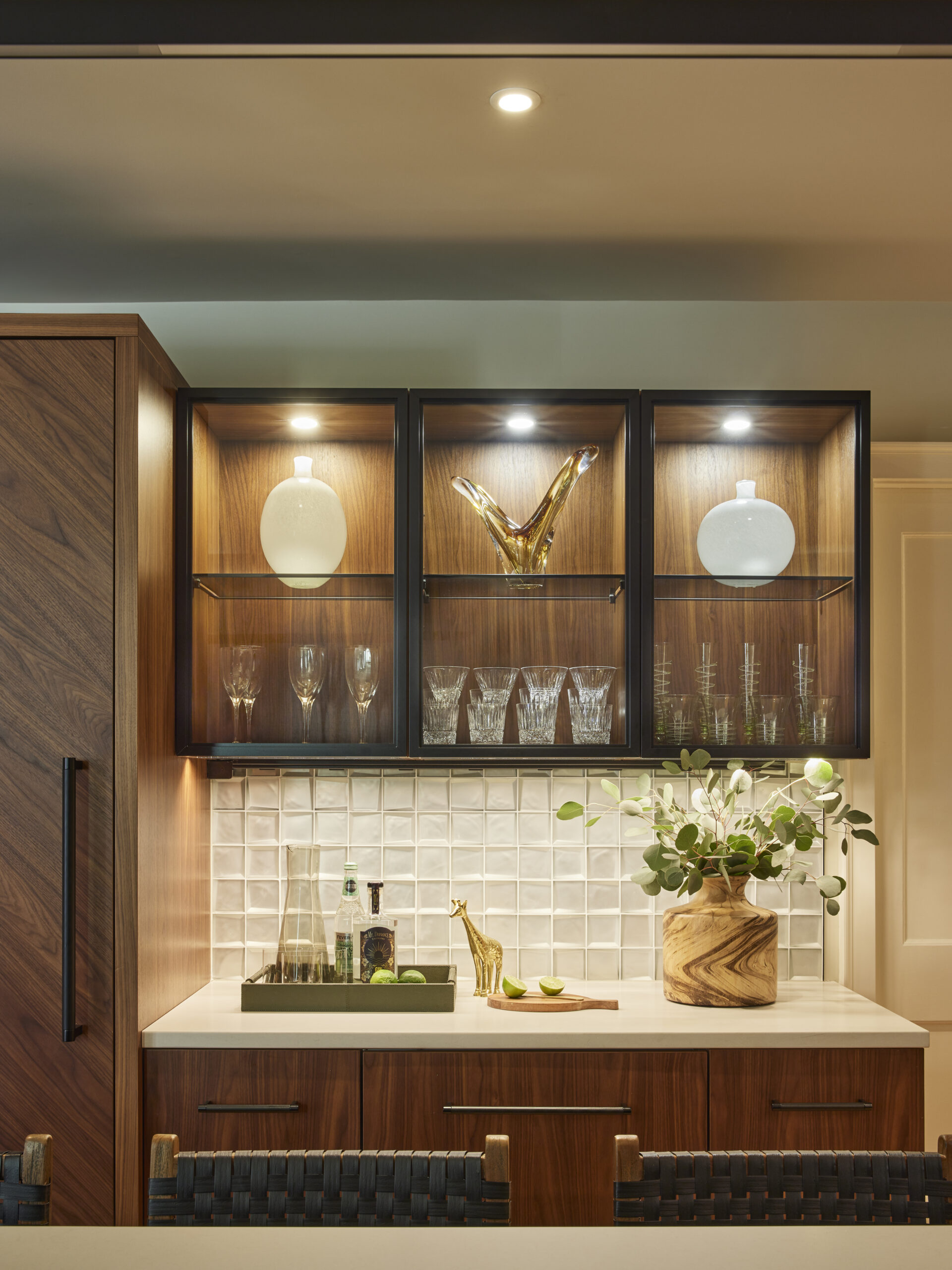 Modern kitchen bar area with wood cabinets, glassware in lit upper shelves, a tray with bottles and glasses, a gold giraffe figurine, and a vase with greenery on a white countertop—an elegant touch of luxury interior design.