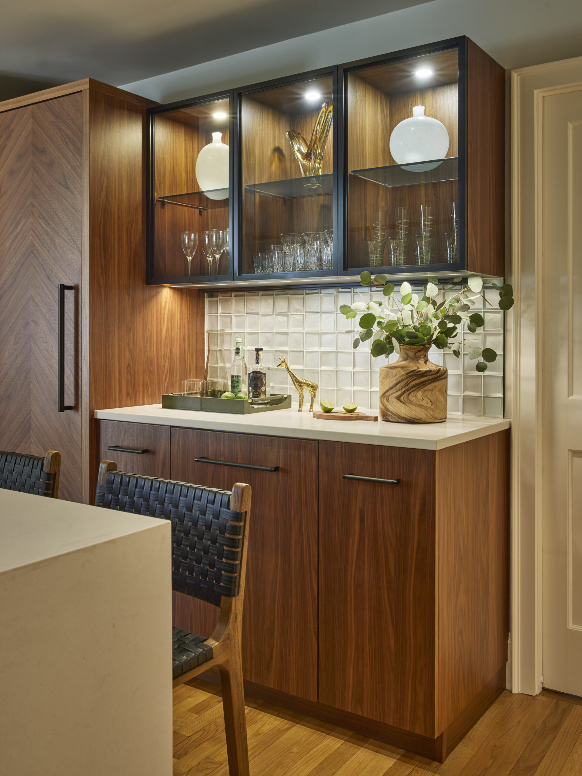 A modern kitchen bar area with wood cabinets, glass display shelves, a white countertop, decorative items, glassware, and a woven-back chair—perfectly embodying luxury interior design by an expert interior designer in Massachusetts.