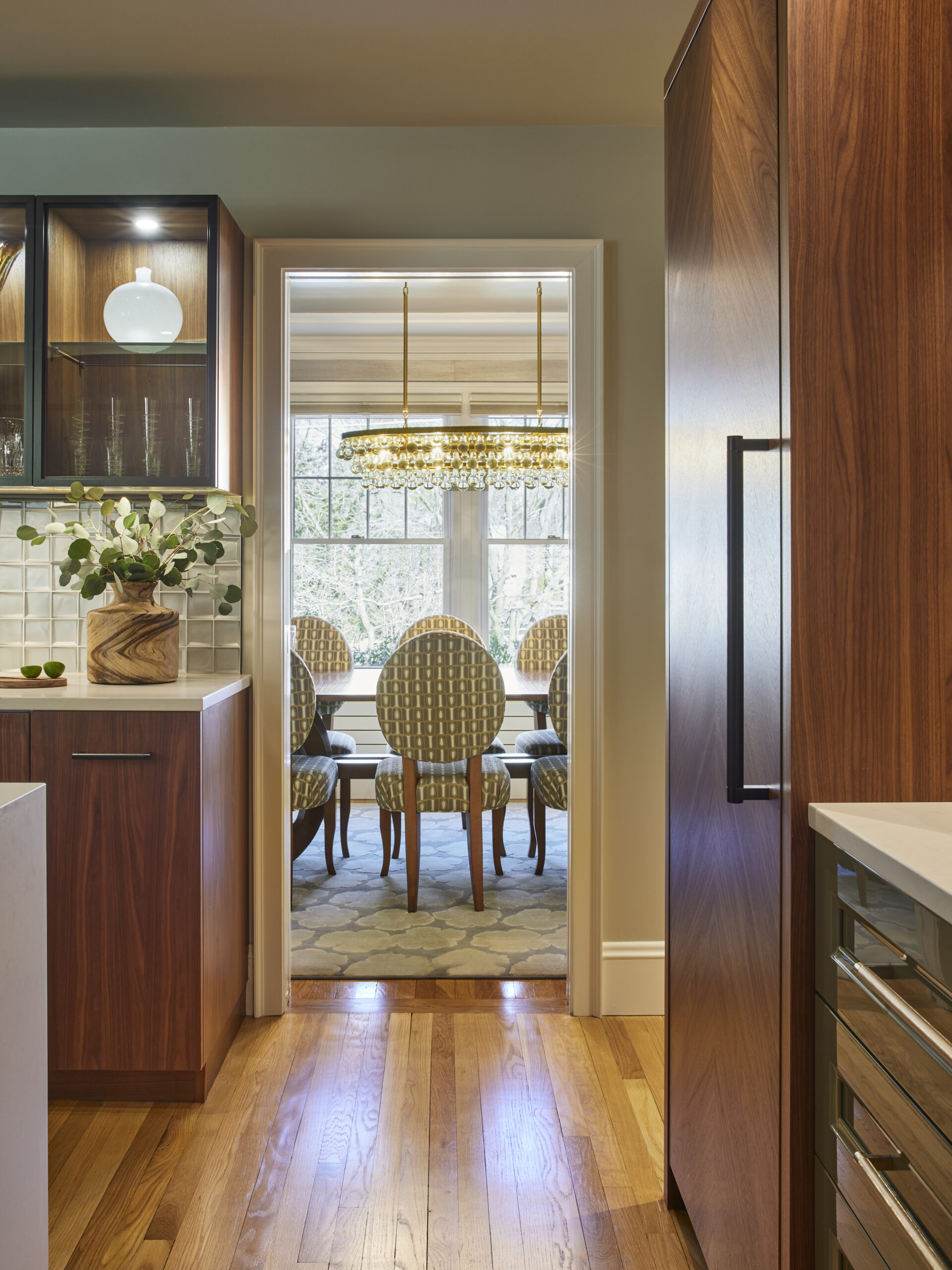 View from a modern kitchen with wooden cabinets into a dining room with patterned chairs and a large chandelier, showcasing luxury interior design by an expert interior designer in Massachusetts.