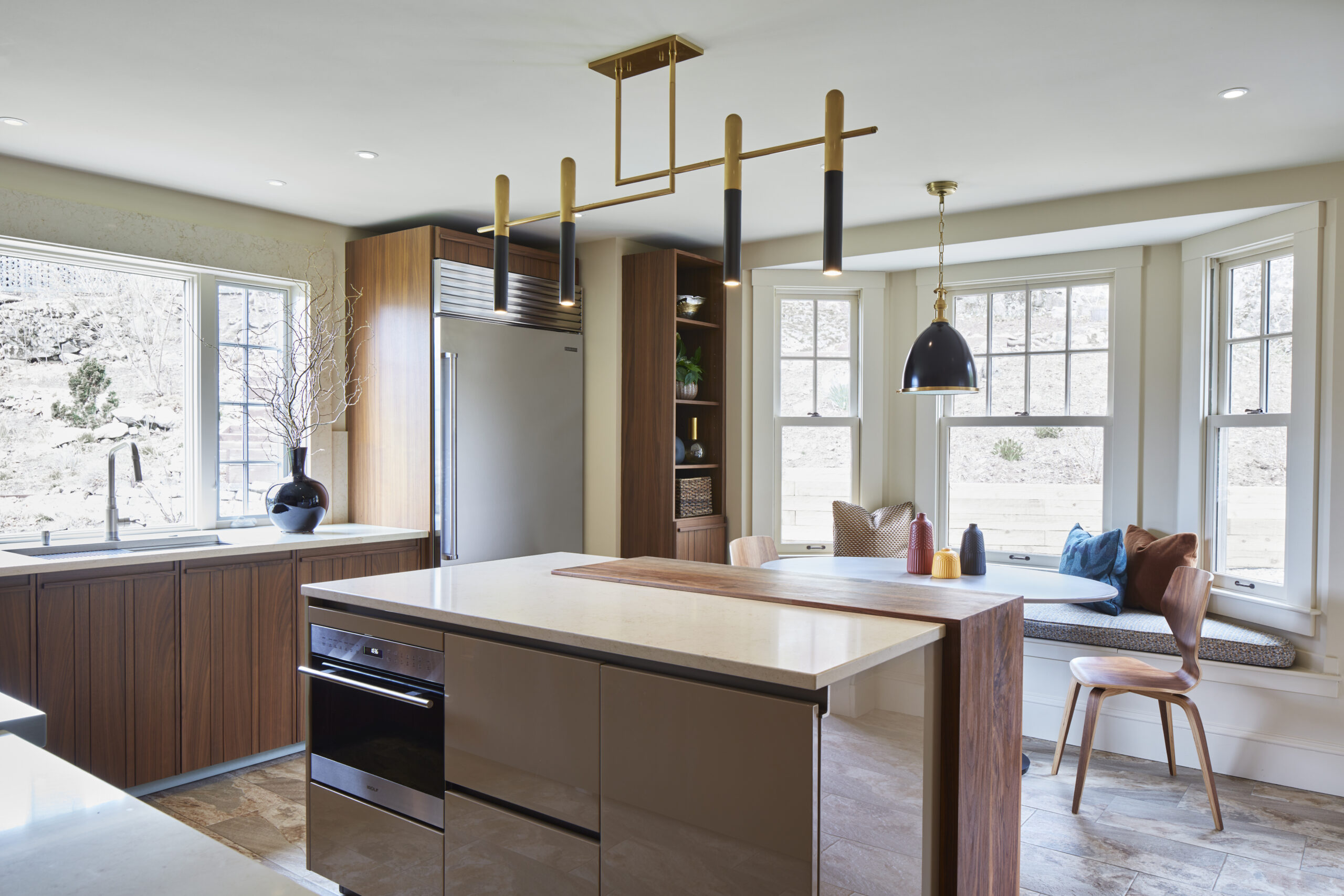 Modern kitchen with wood cabinetry, an island with built-in oven, stainless steel appliances, large windows, and a cozy breakfast nook—exemplifying luxury interior design by a skilled interior designer in Massachusetts.